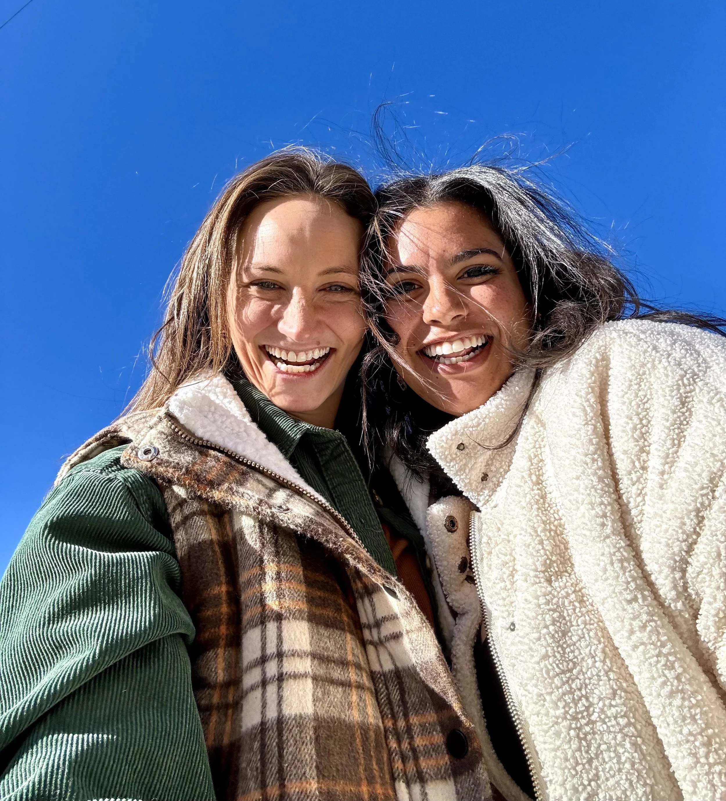 Two smiling women taking a selfie outdoors against a bright blue sky, wearing warm jackets.