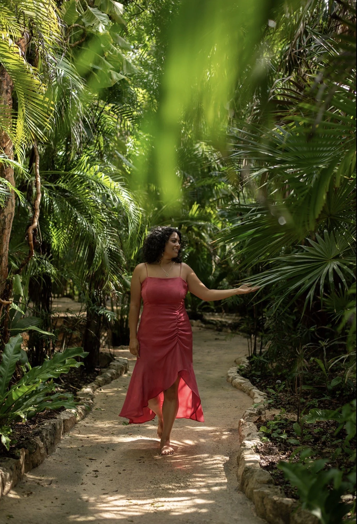 A woman in a pink dress walking barefoot on a garden pathway surrounded by lush green tropical plants.