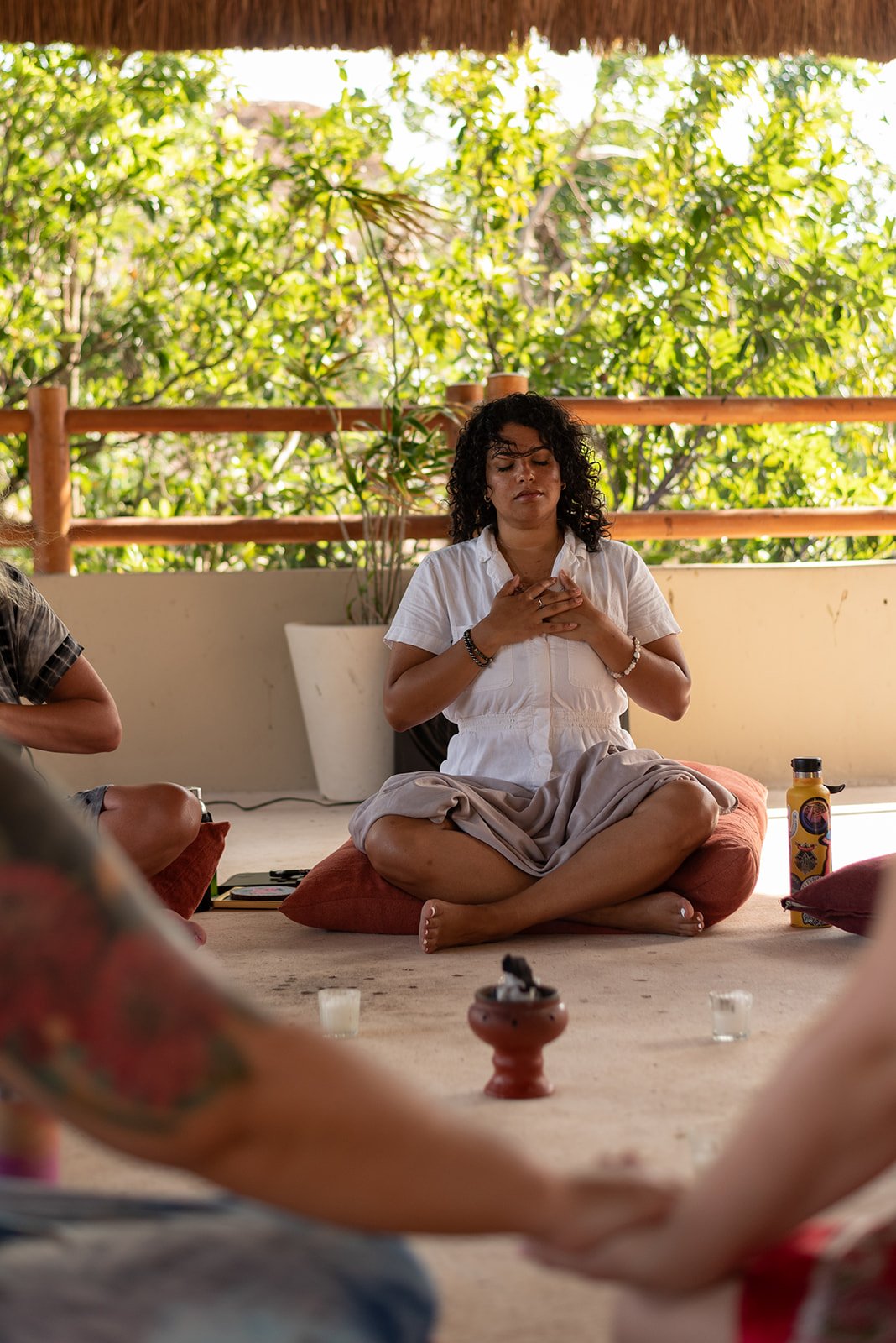 A woman sitting cross-legged on a cushion, with her hands on her chest, during a meditation session surrounded by others, with a natural outdoor setting in the background.