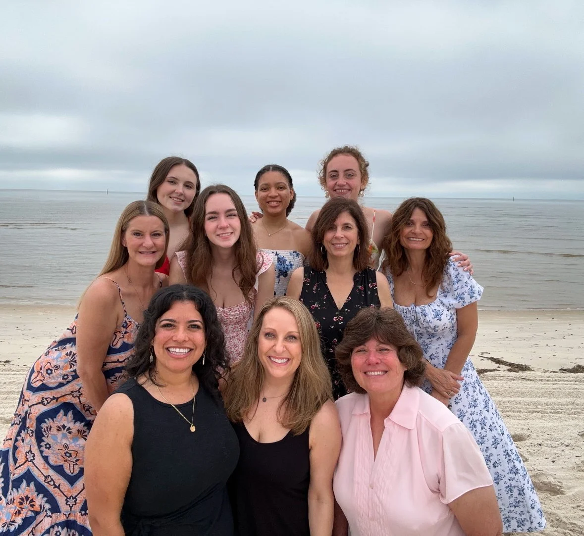 Group of ten women smiling on a beach with cloudy sky and ocean in background.