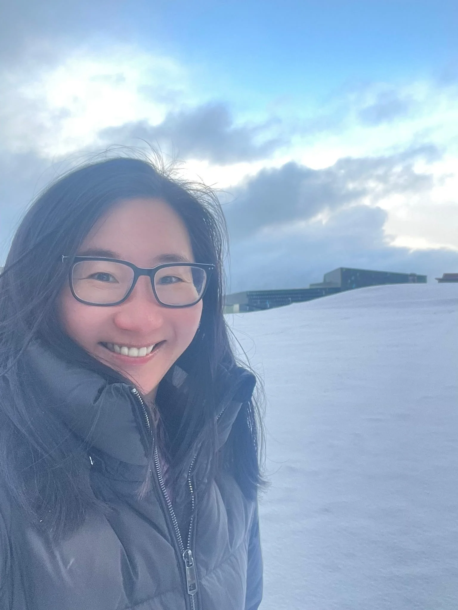 A smiling woman wearing glasses and a dark jacket taking a selfie in a snowy outdoor landscape with a building in the distance and cloudy sky overhead.