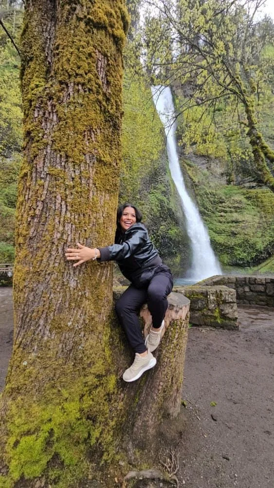 Woman smiling and hugging a large mossy tree trunk near a waterfall in a lush forest.