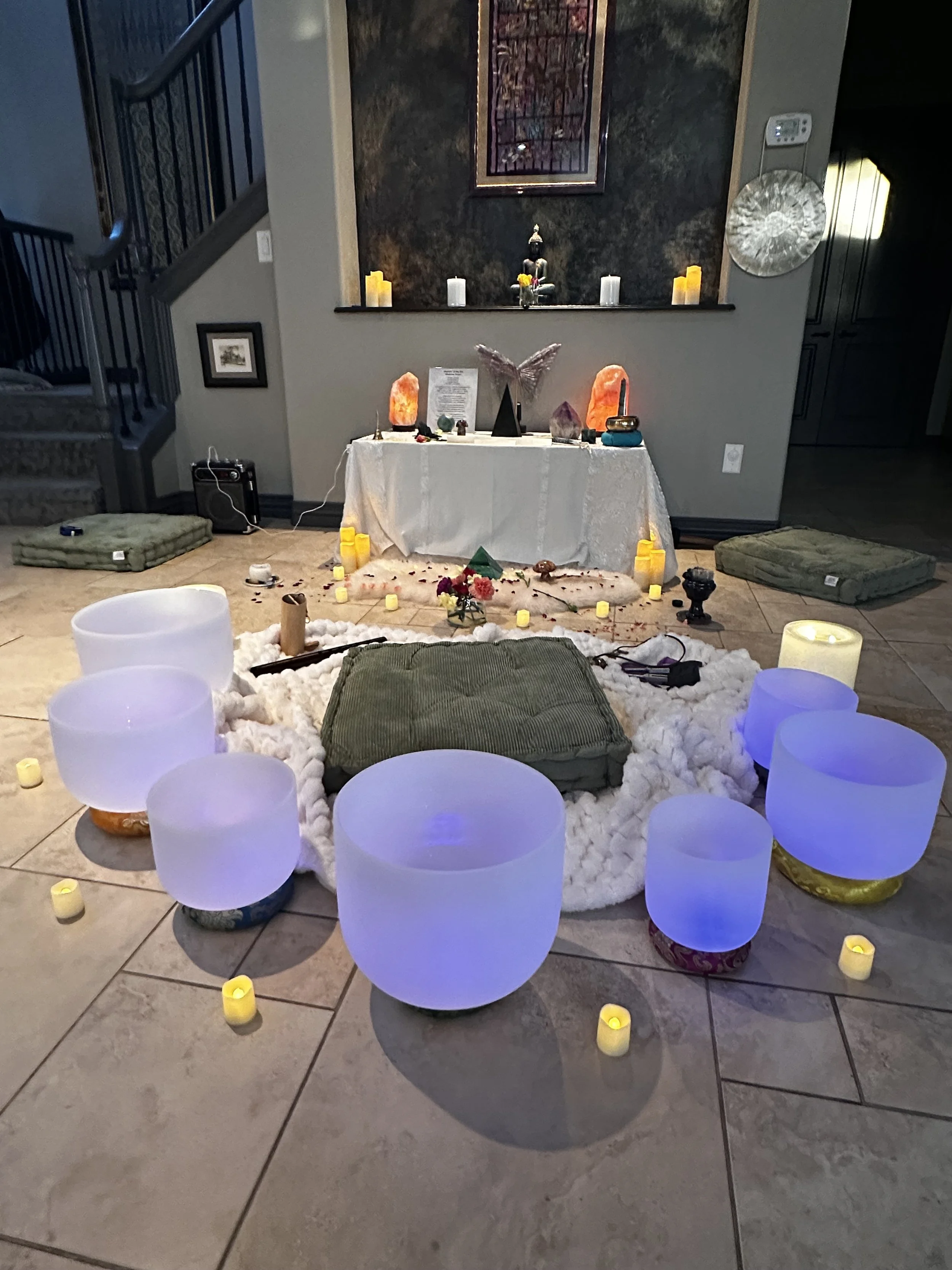 A cozy indoor setting with a white table draped with a cloth, surrounded by soft glowing LED candles and cushions on the floor. A small altar or meditation space with statues, salt lamps, crystals, and flowers is set up on the table. The background features candles and decorations on a ledge against a dark wall, with framed artwork and wall decor. The atmosphere is calm and spiritual, with dim lighting and decorative elements.