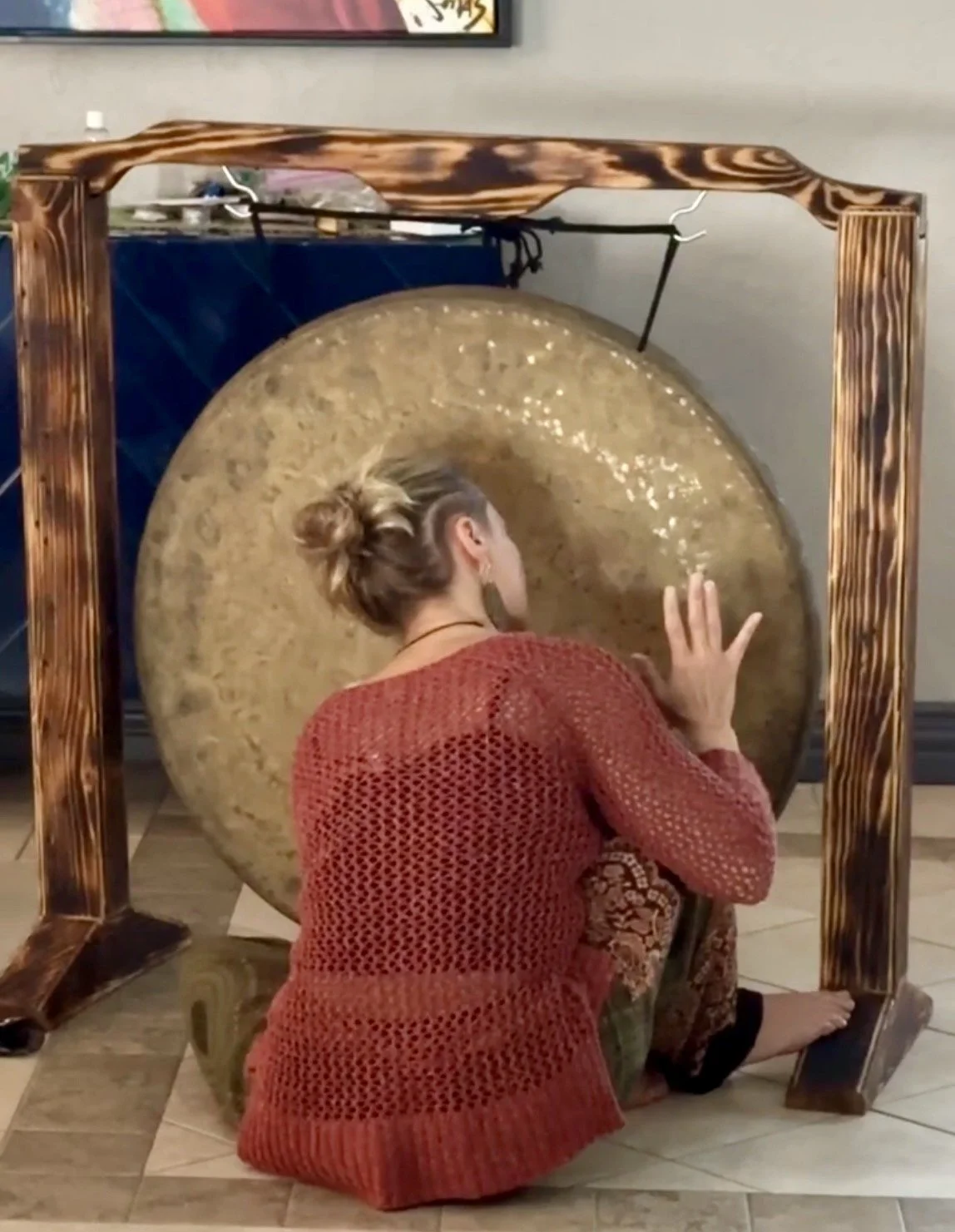 A woman with blonde hair in a bun and wearing a red knitted sweater is kneeling and touching a large gong with her hand, in an indoor setting.