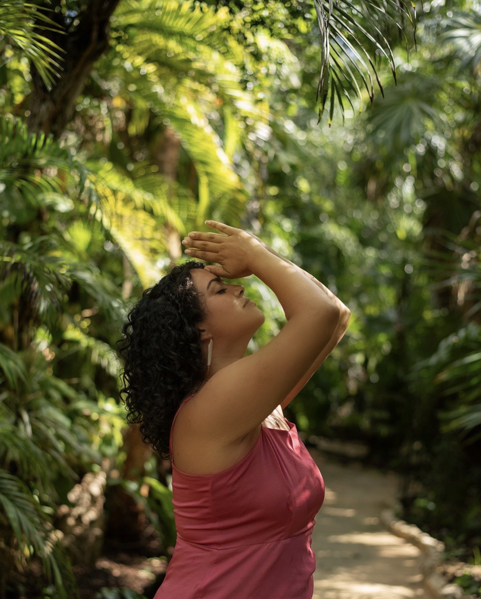 A woman with curly dark hair and medium skin tone is standing outdoors in a lush, green, tropical environment, with her eyes closed and one hand raised to her forehead in a relaxed pose.