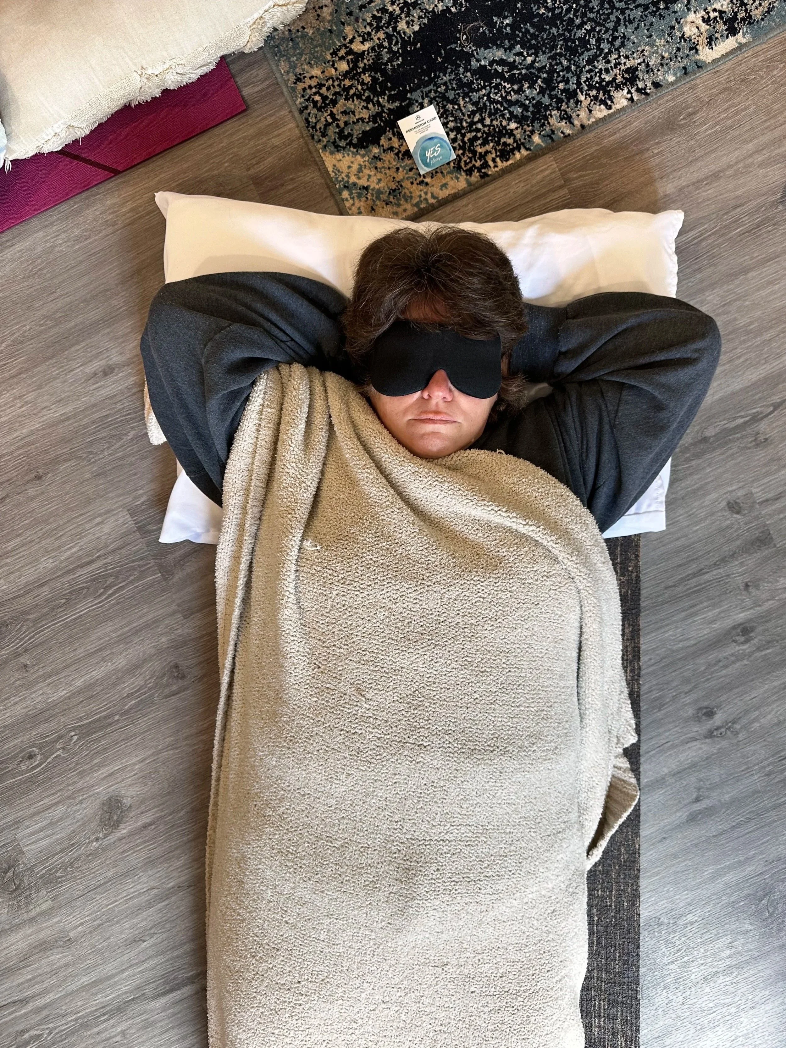 Person lying in bed with eyes covered by a sleep mask, covered with a blanket, on a bed with a pillow, next to a wooden floor and a patterned rug, with a card on the rug.