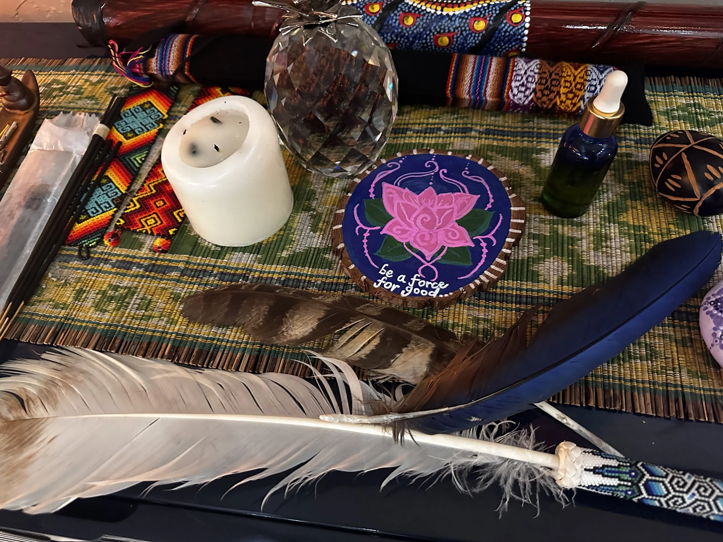 A collection of spiritual and decorative items, including feathers, a beaded beaded textile item, a white candle, a large faceted crystal, and a painted wooden item with a pink lotus flower and the words "be a force for good" on a colorful woven surface.