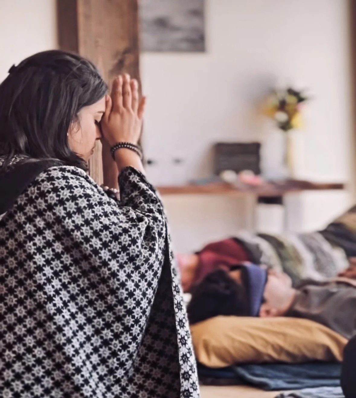 A woman praying with her hands pressed against a wall, eyes closed, while a person lies motionless on a bed in the background.
