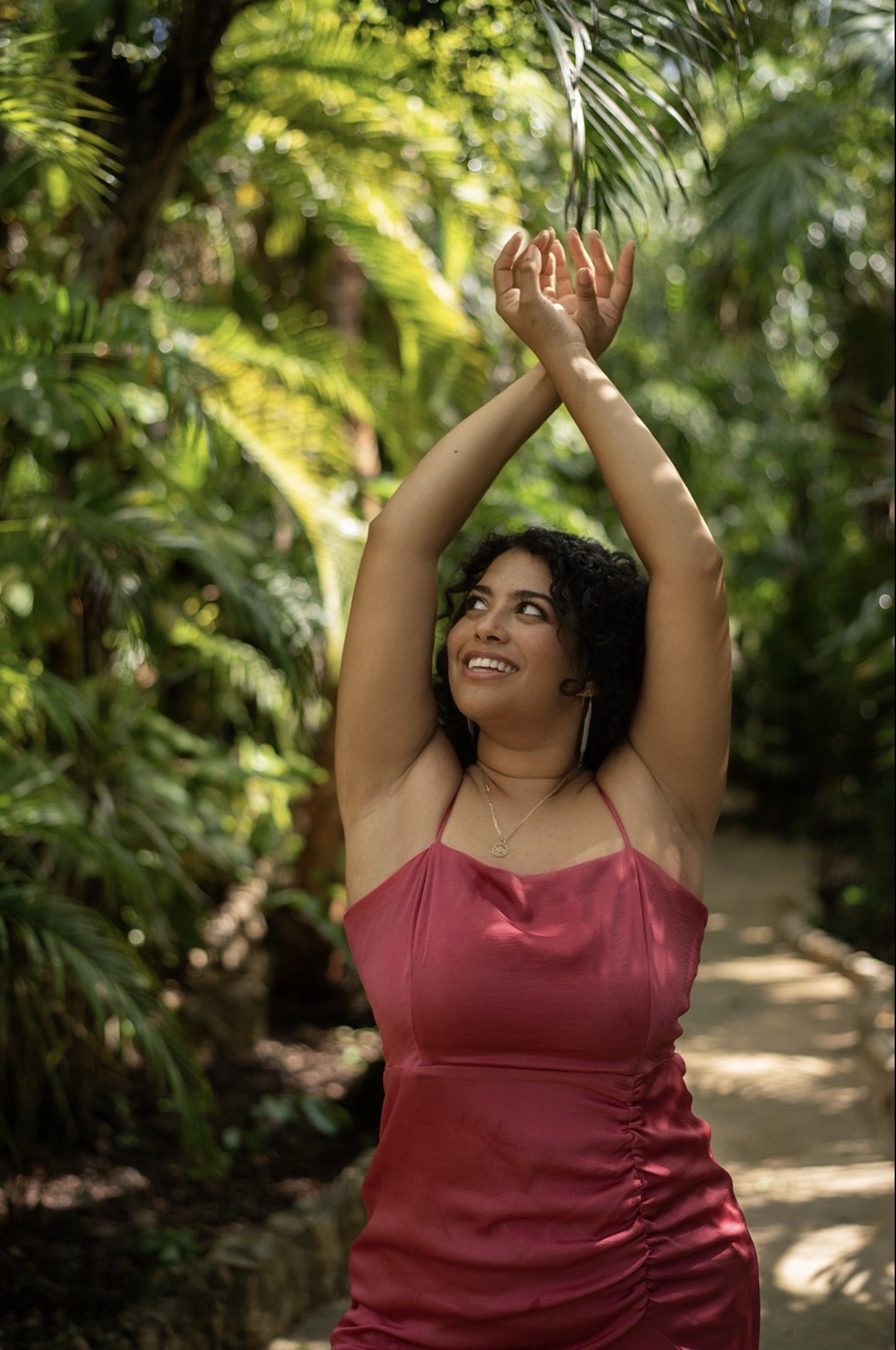 A woman with curly dark hair smiling and looking up, standing outdoors in a lush green tropical setting, wearing a red satin dress with spaghetti straps, her arms raised above her head with hands crossed at the wrists.