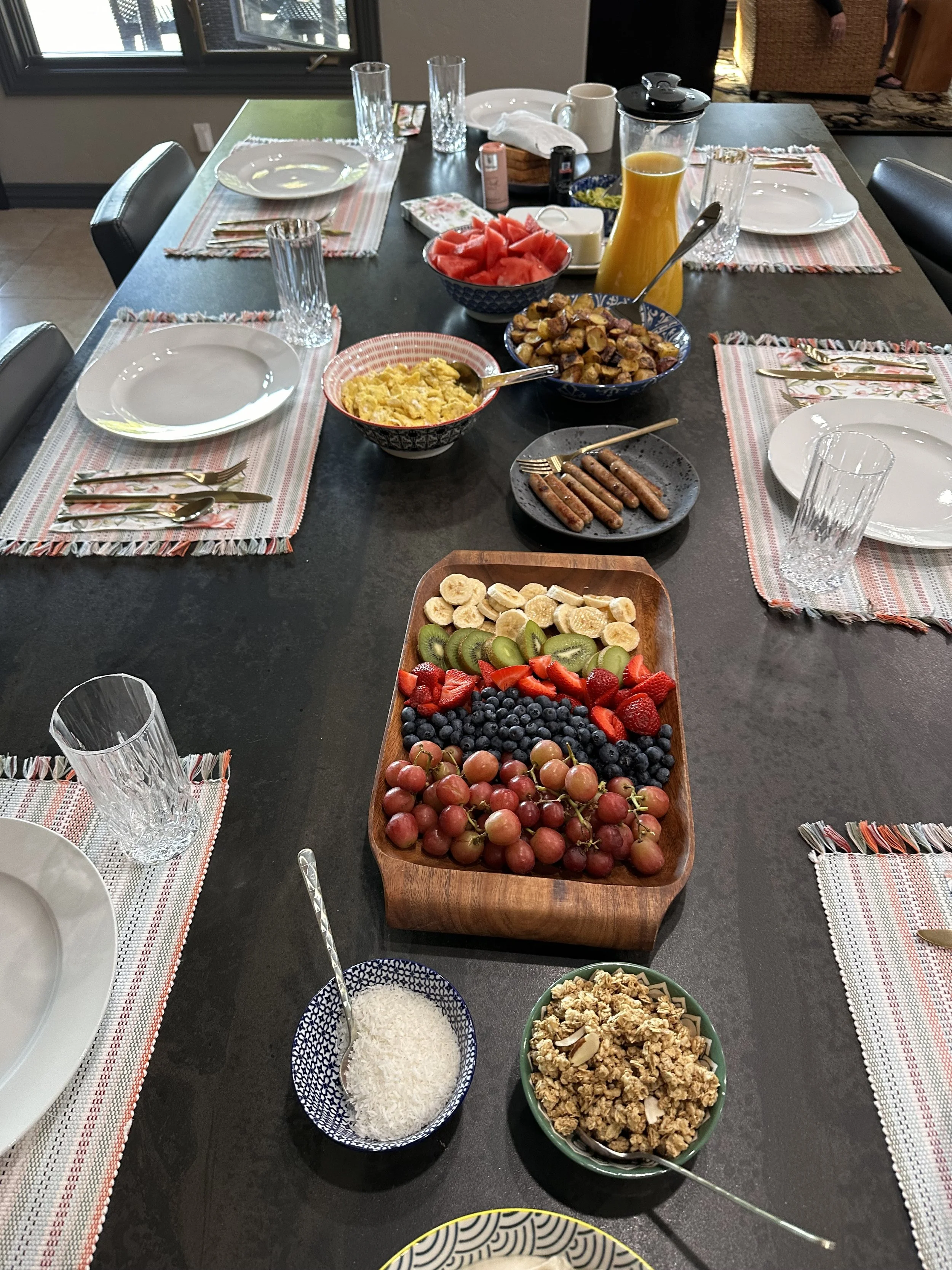 A breakfast or brunch table set with plates, glasses, utensils, and colorful bowls filled with fresh fruit, scrambled eggs, breakfast sausage, and granola. There is also a pitcher of orange juice on the table.
