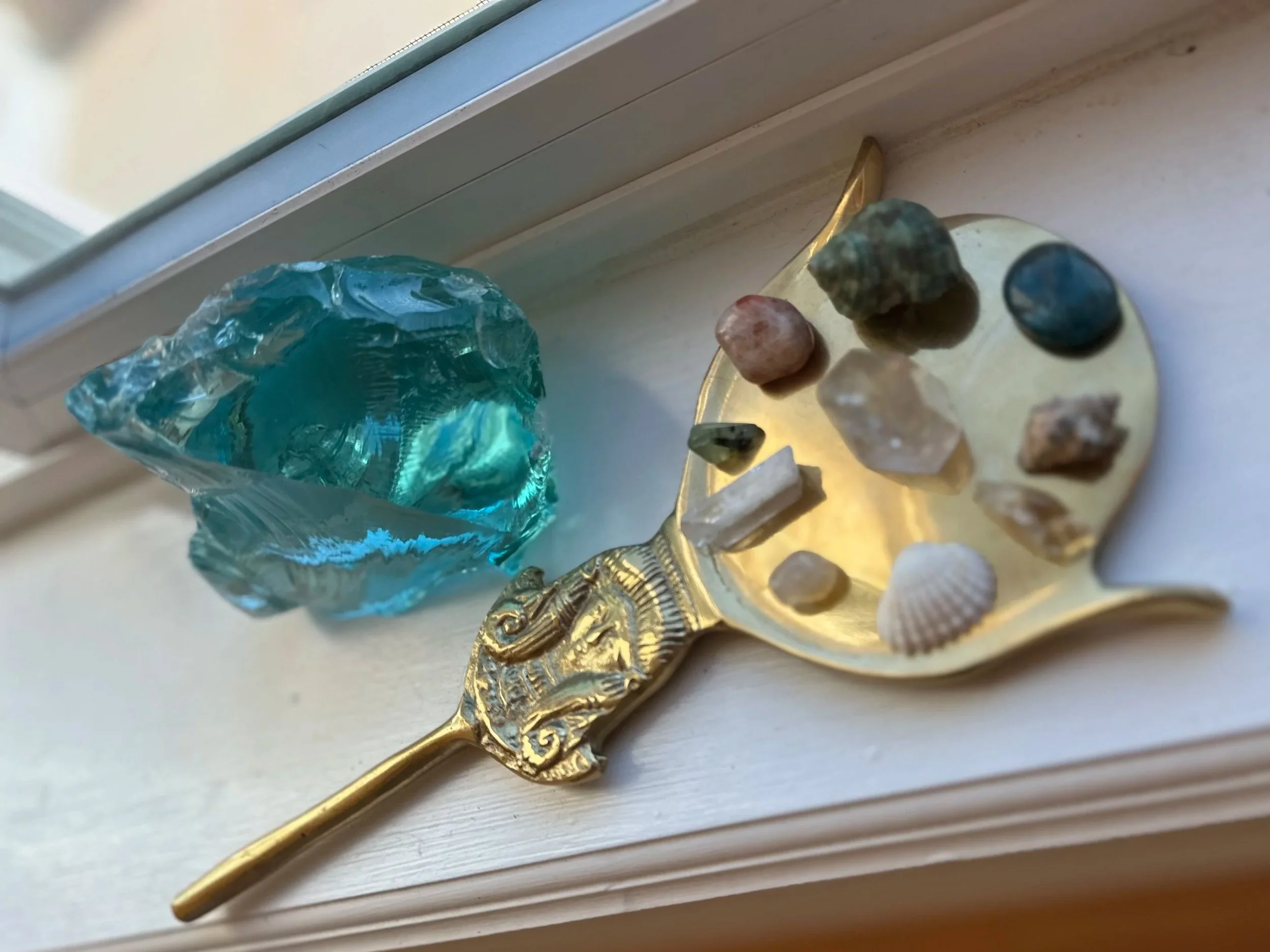 Decorative gold-colored metal key with an ornate handle, lying on a white wooden surface. Next to the key is a collection of small gemstones and a large piece of turquoise-colored glass or stone. The background shows part of a window frame or ledge.