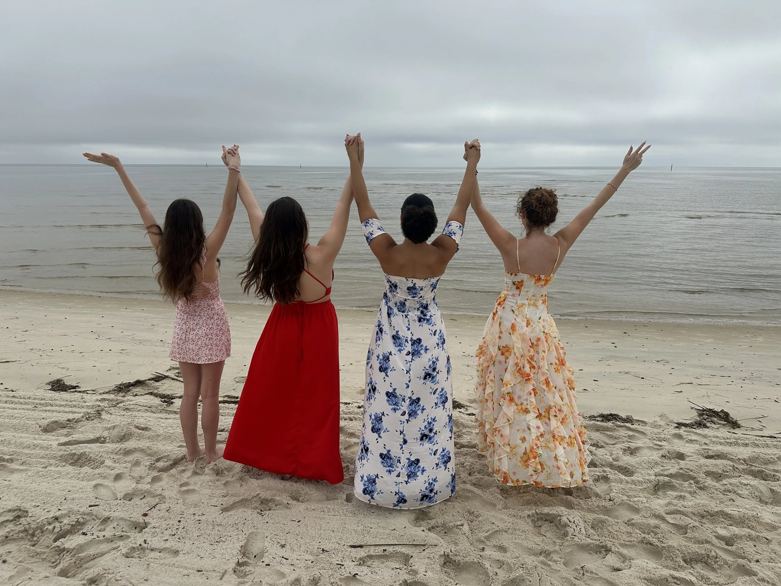 Four women standing on a sandy beach with their backs to the camera, holding hands and raising their arms in celebration, with overcast sky and calm ocean in the background.