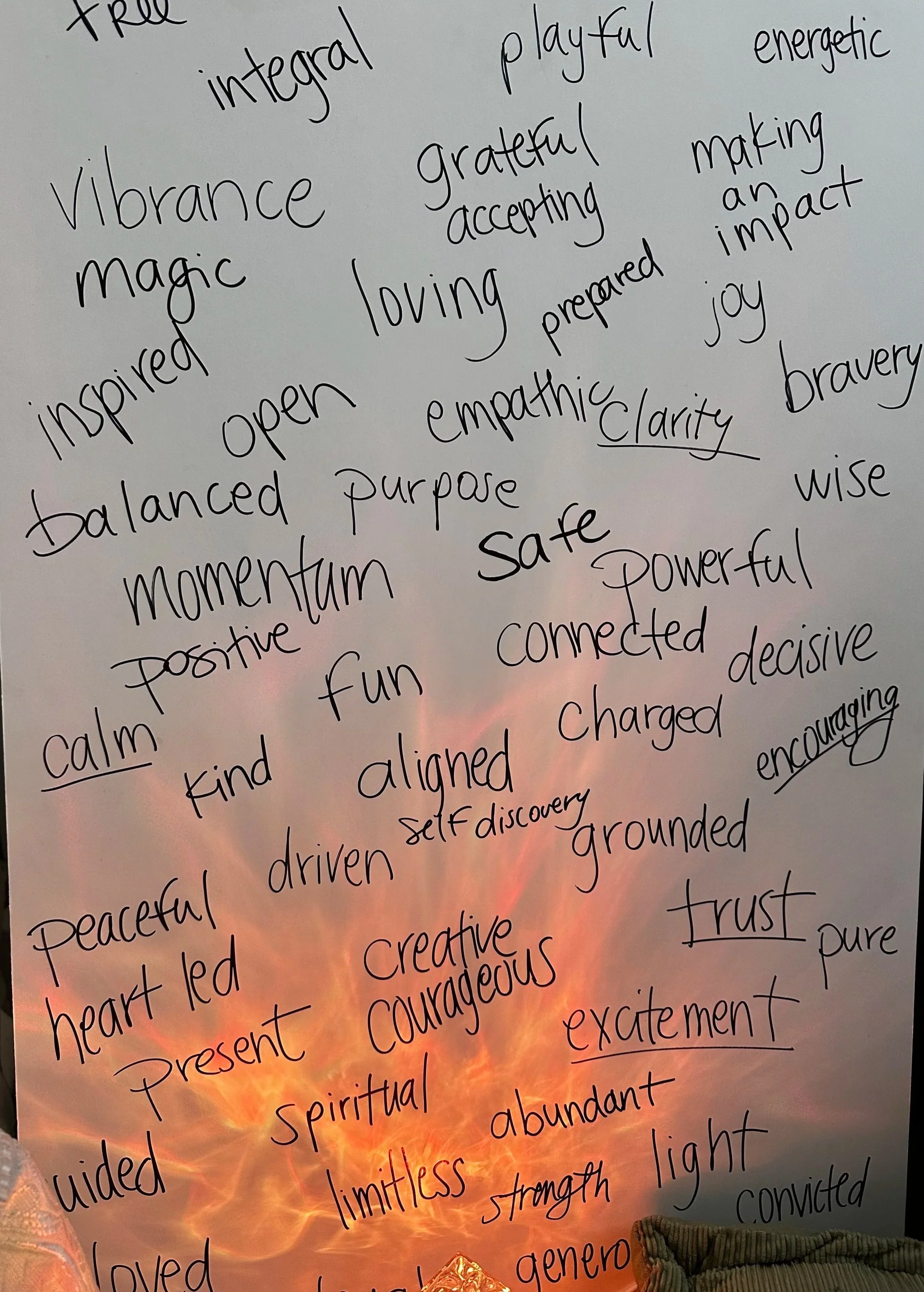 A whiteboard filled with various positive words and phrases written in black marker. The words include 'truth,' 'integral,' 'playful,' 'energetic,' 'vibrance,' 'grateful,' 'accepting,' 'making an impact,' 'magic,' 'loving,' 'inspired,' 'open,' 'prepared,' 'joy,' 'balanced,' 'empathic,' 'caring,' 'bravery,' 'mindful,' 'calm,' 'positive,' 'aligned,' 'self discovery,' 'grounded,' 'driven,' 'focused,' 'heart led,' 'present,' 'powerful,' 'creative,' 'courageous,' 'limitless,' 'light,' and many others. The background shows a sunset with warm orange and pink hues.