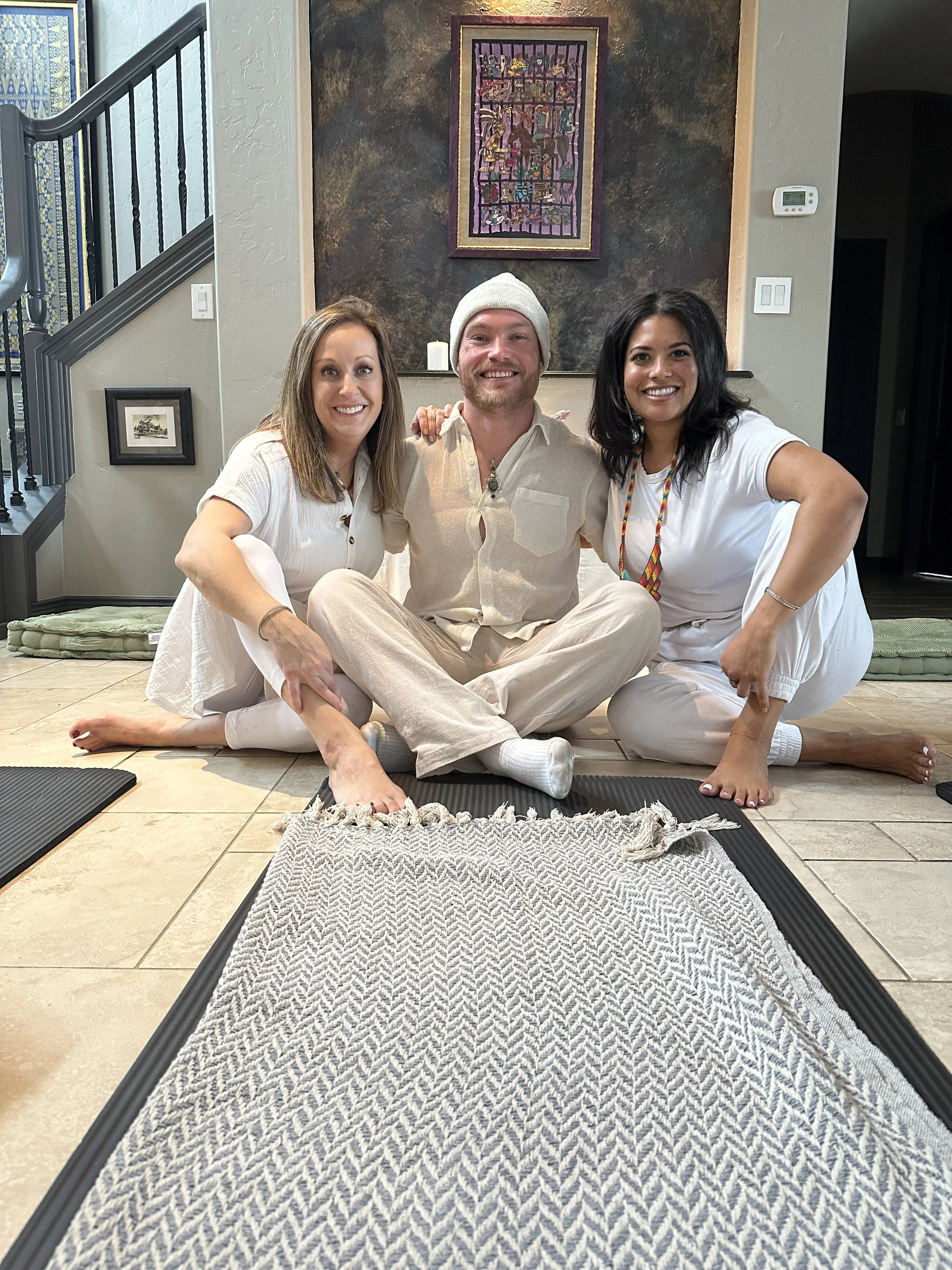 Three people sitting on the floor inside a house, smiling at the camera. The person in the middle is male, wearing a white beanie, beige shirt, and beige pants. The two women on either side are dressed in white, with the woman on the left having light brown hair, and the woman on the right having dark hair and wearing a necklace. They are near yoga mats with a textured gray and white rug in the foreground, with a painted wall and framed artwork behind them.