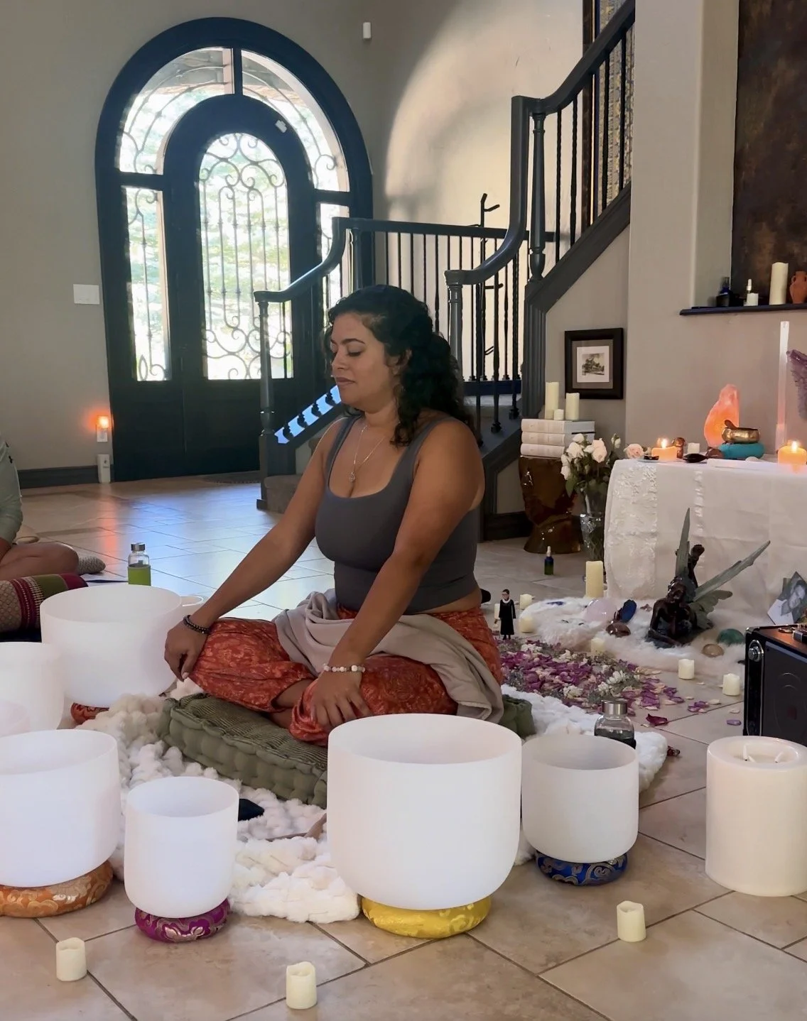 A woman sitting cross-legged on the floor during a meditation or spiritual ceremony, surrounded by singing bowls, candles, and spiritual objects in a cozy room with an arched glass door in the background.
