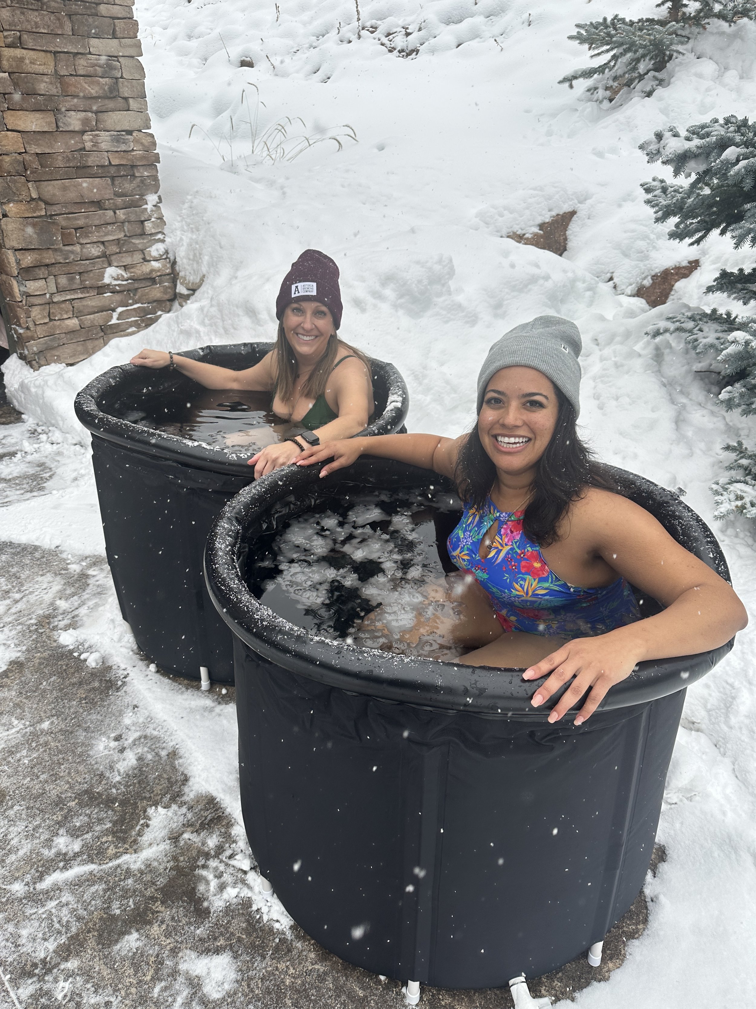 Two women sitting in black outdoor hot tubs outside in a snowy environment, smiling and wearing beanies.