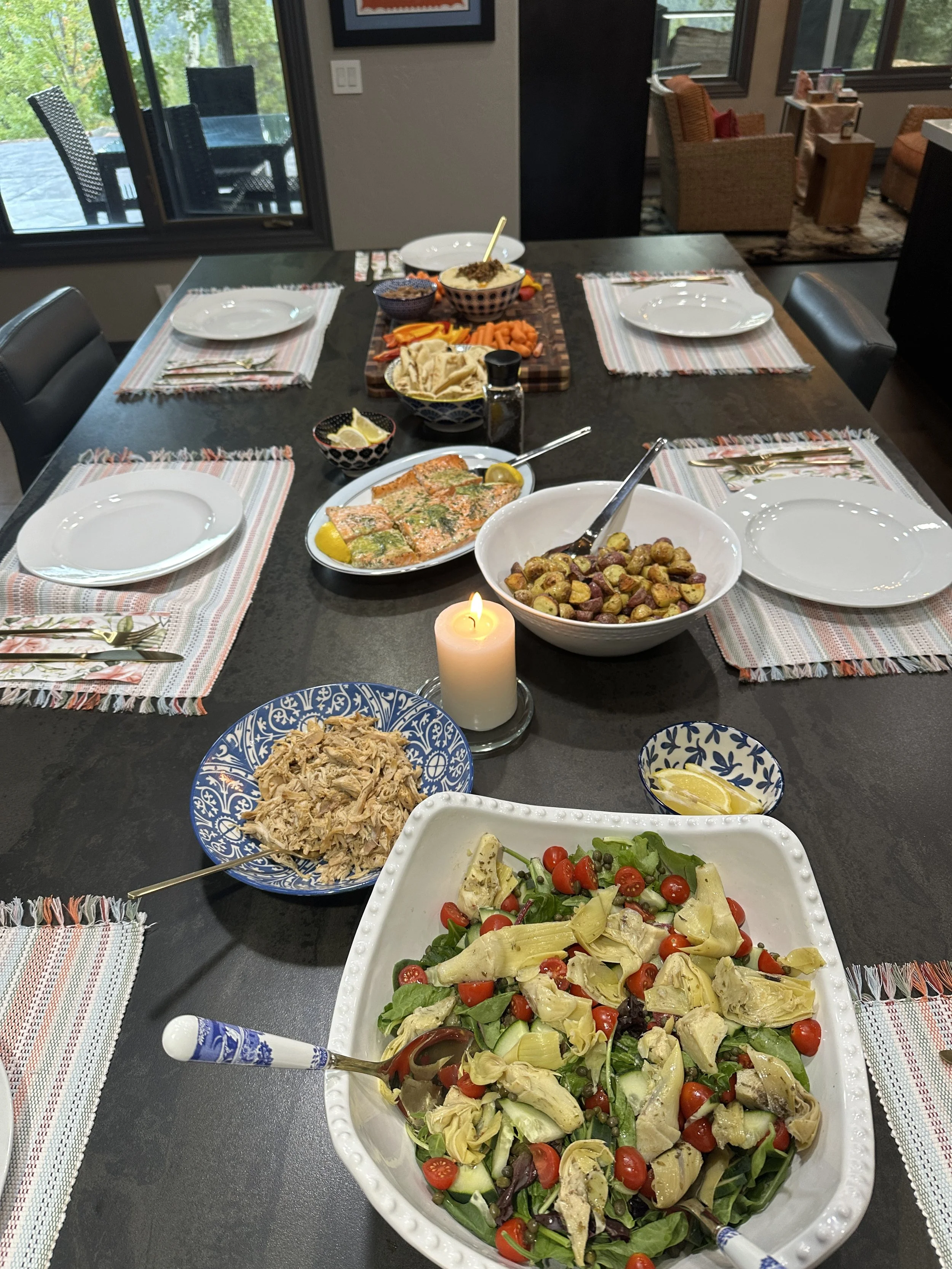 A dining table set with plates, bowls of salad, a bowl of pasta, a bowl of roasted vegetables, and a candle. In the background, there are chairs and a patio outside the window.