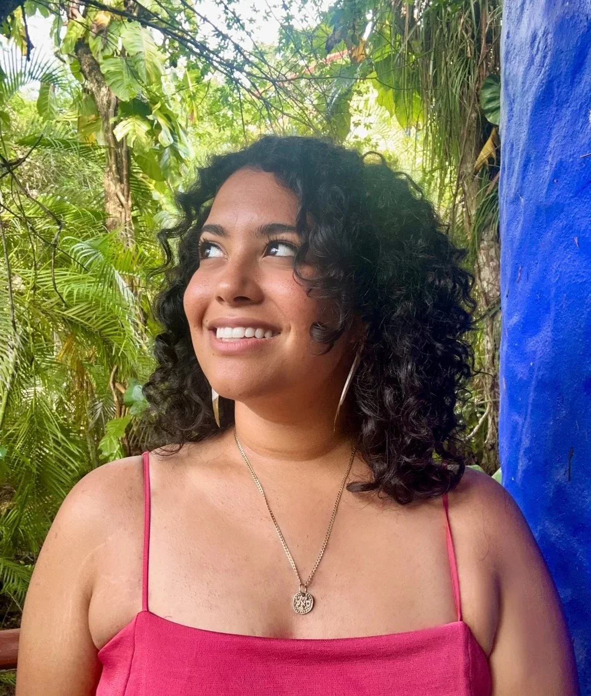 A woman with curly dark hair wearing a pink spaghetti strap top and silver jewelry, standing outdoors in a lush green environment with a blue painted wall on her right side, smiling and looking slightly upwards.
