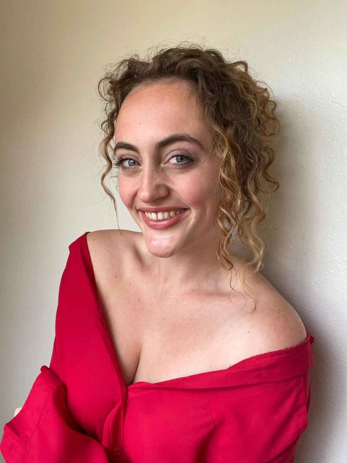 A woman with curly hair wearing a red off-the-shoulder top, smiling and standing against a plain wall.