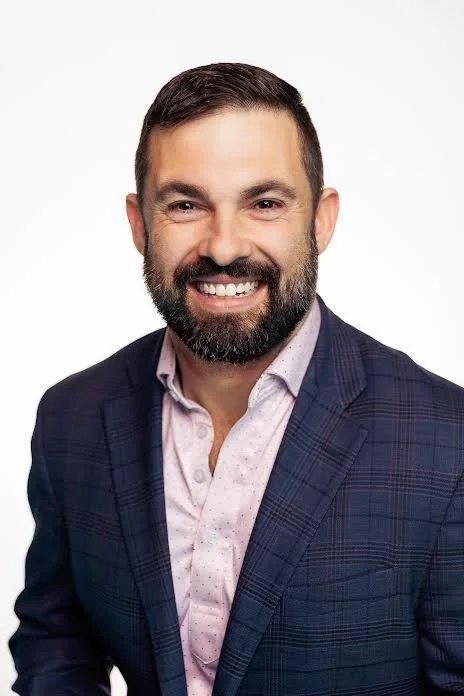 Headshot of a smiling man with dark hair, a beard, wearing a dark blue checkered blazer and a light pink button-up shirt against a white background.