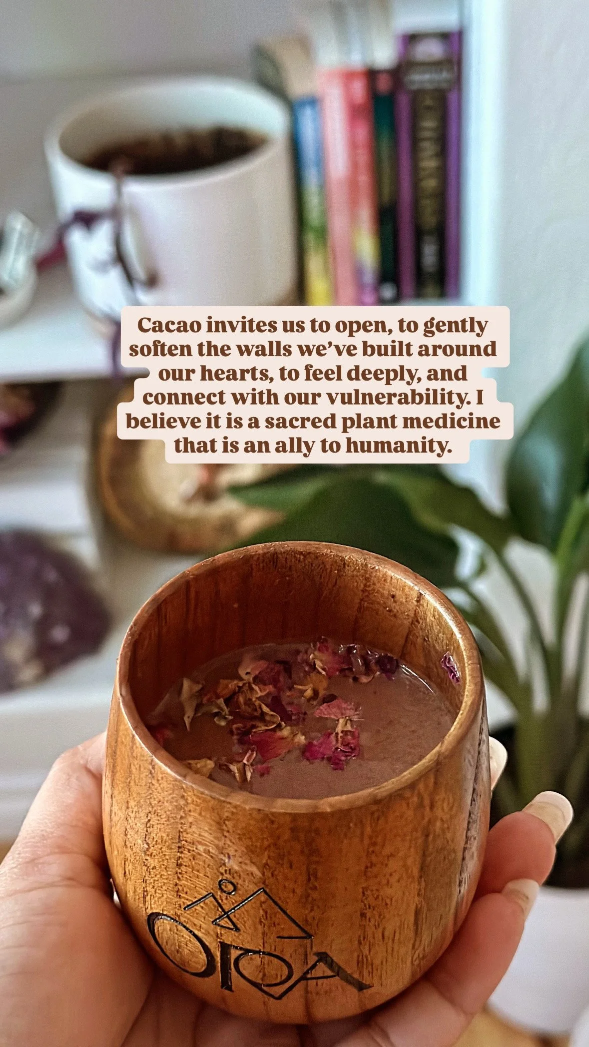 A wooden cup or mug filled with tea, topped with dried flower petals, being held in hand. In the background, a blurred white shelf with a potted plant, colorful books, and a white container with a spoon.