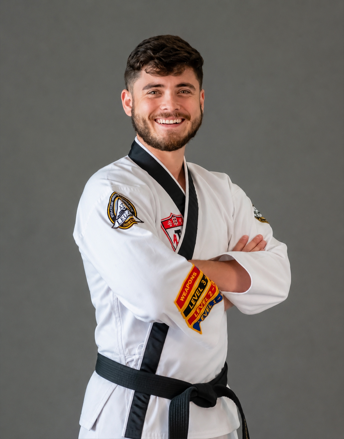 A smiling man wearing a white taekwondo uniform and a black belt, standing with arms crossed against a gray background.