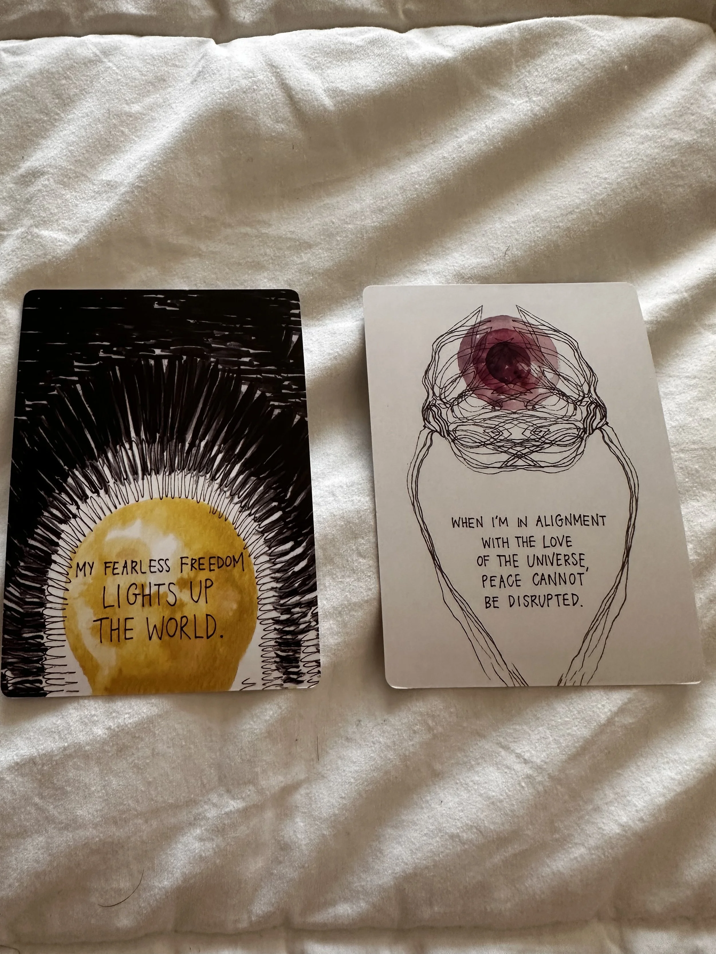 Two tarot cards on a soft, light-colored fabric surface. The card on the left features a yellow sun with rays and the phrase, "My fearless freedom lights up the world." The card on the right depicts an abstract line drawing of a human skull with a purple-red watercolor circle on top, and the quote, "When I'm in alignment with the love of the universe peace cannot be disrupted."