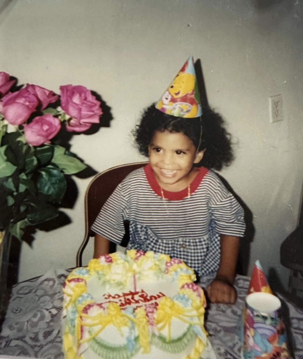 A young girl celebrating her birthday, wearing a birthday hat with Winnie the Pooh on it, smiling, sitting at a table with a colorful birthday cake, pink roses on the side, and party cups, with a wall in the background.