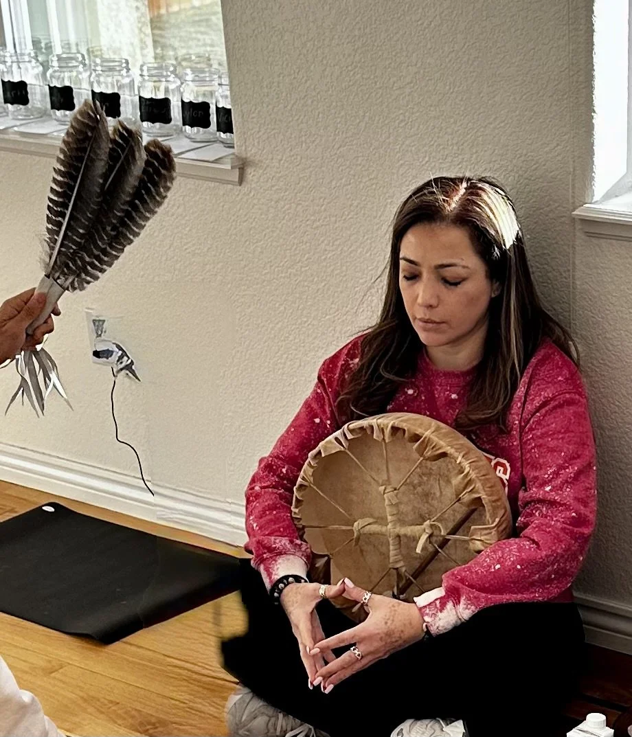 A woman with long dark hair, wearing a red sweatshirt, sitting on the floor against a wall, holding a circular object wrapped in paper and string, with feathers held by a person standing nearby. There are glass jars on a shelf behind her.
