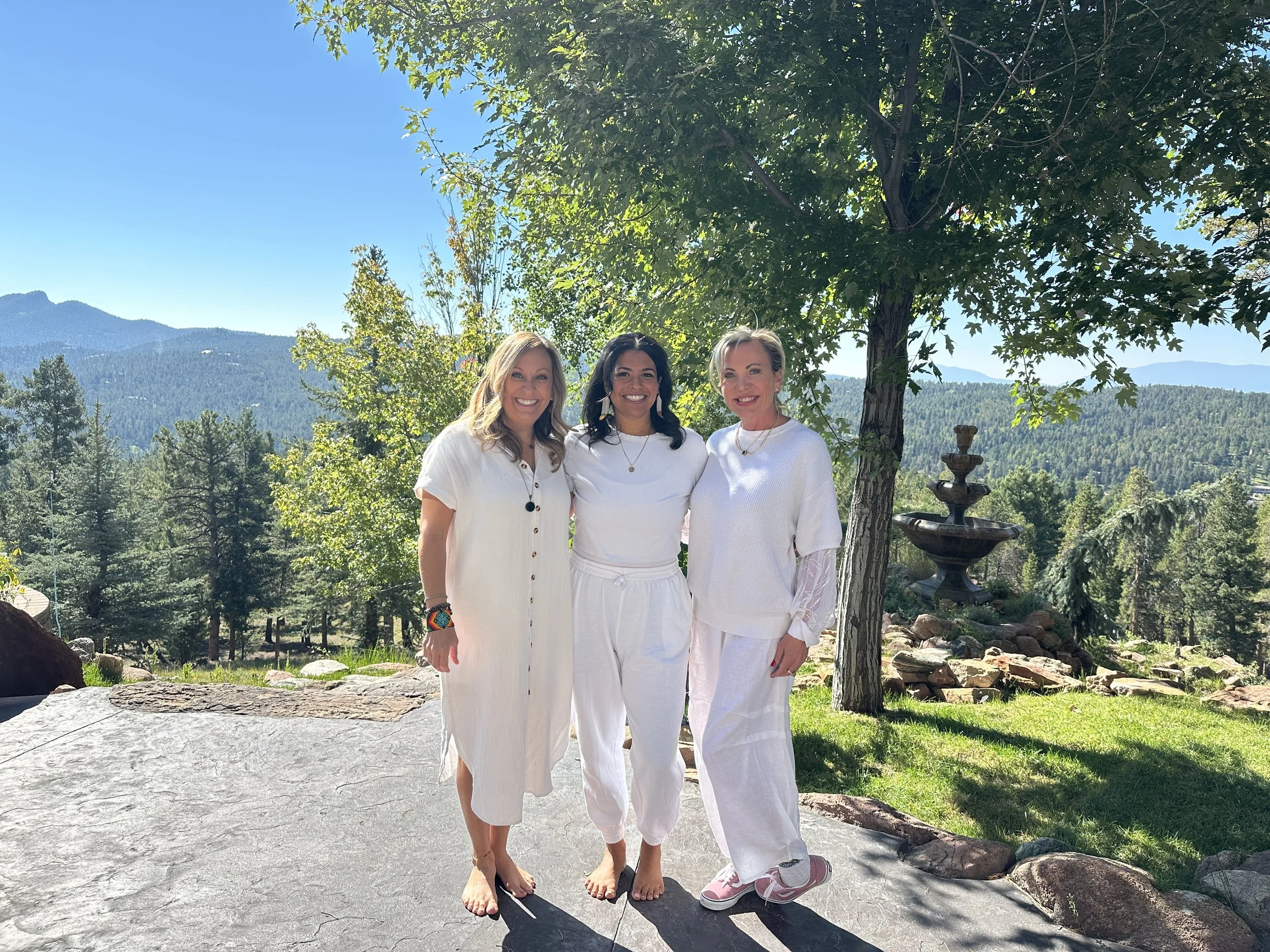 Three women standing outdoors on a sunny day, dressed in white, with mountains, trees, and a fountain in the background.