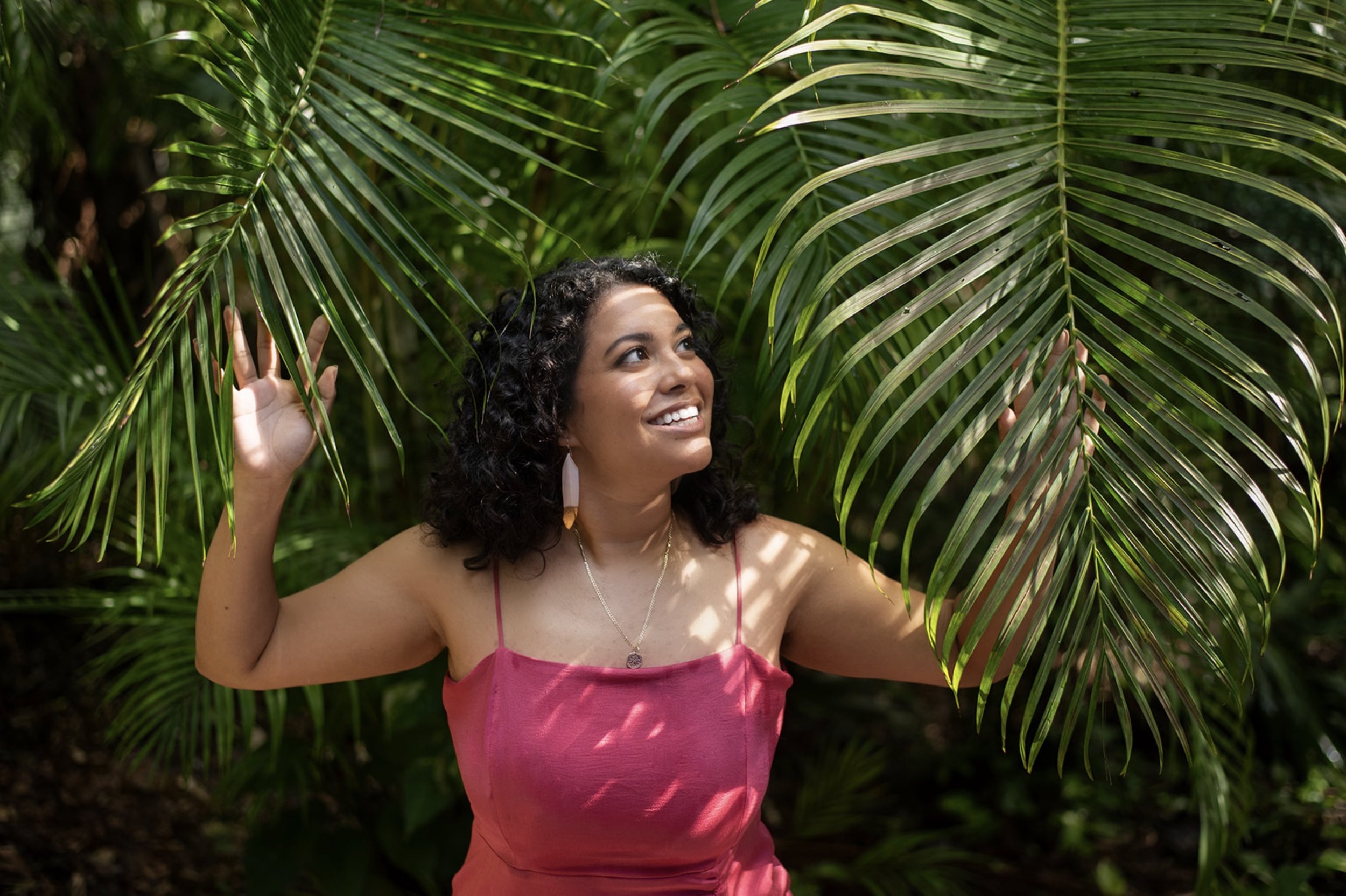 A woman with curly dark hair smiling and wearing a pink dress, standing among lush green palm leaves in a tropical setting.