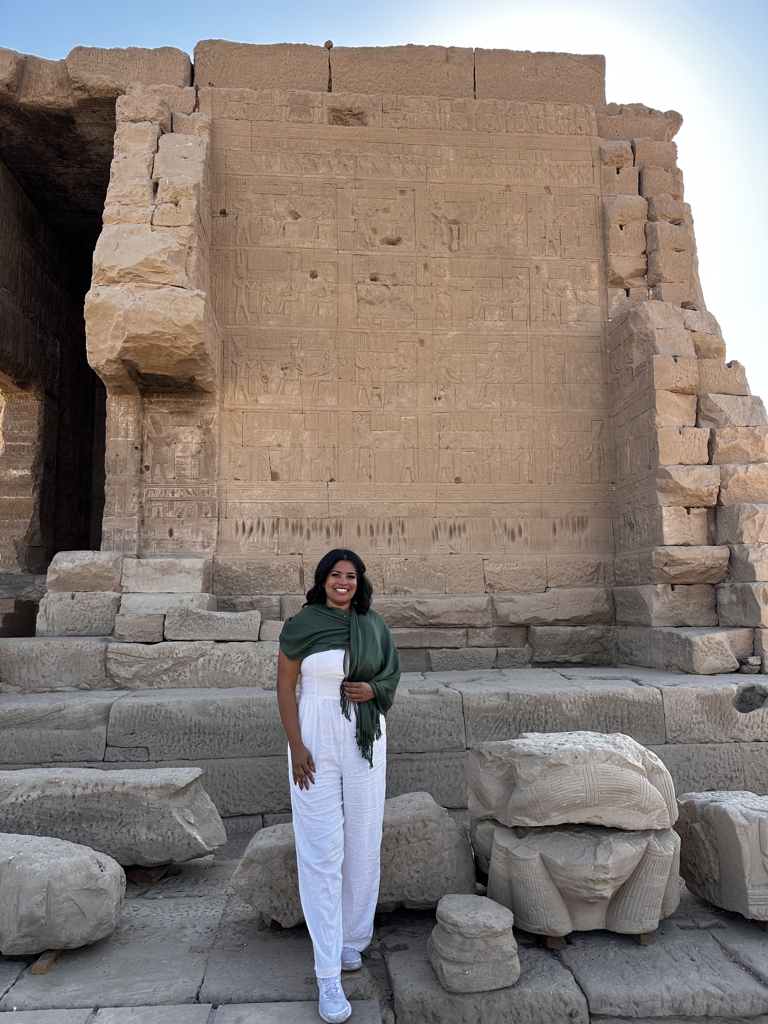 A smiling woman dressed in white pants and a green shawl stands in front of an ancient stone temple wall with carved hieroglyphics. The structure is made of large stone blocks, with some fragments of sculptures on the ground surrounding her.