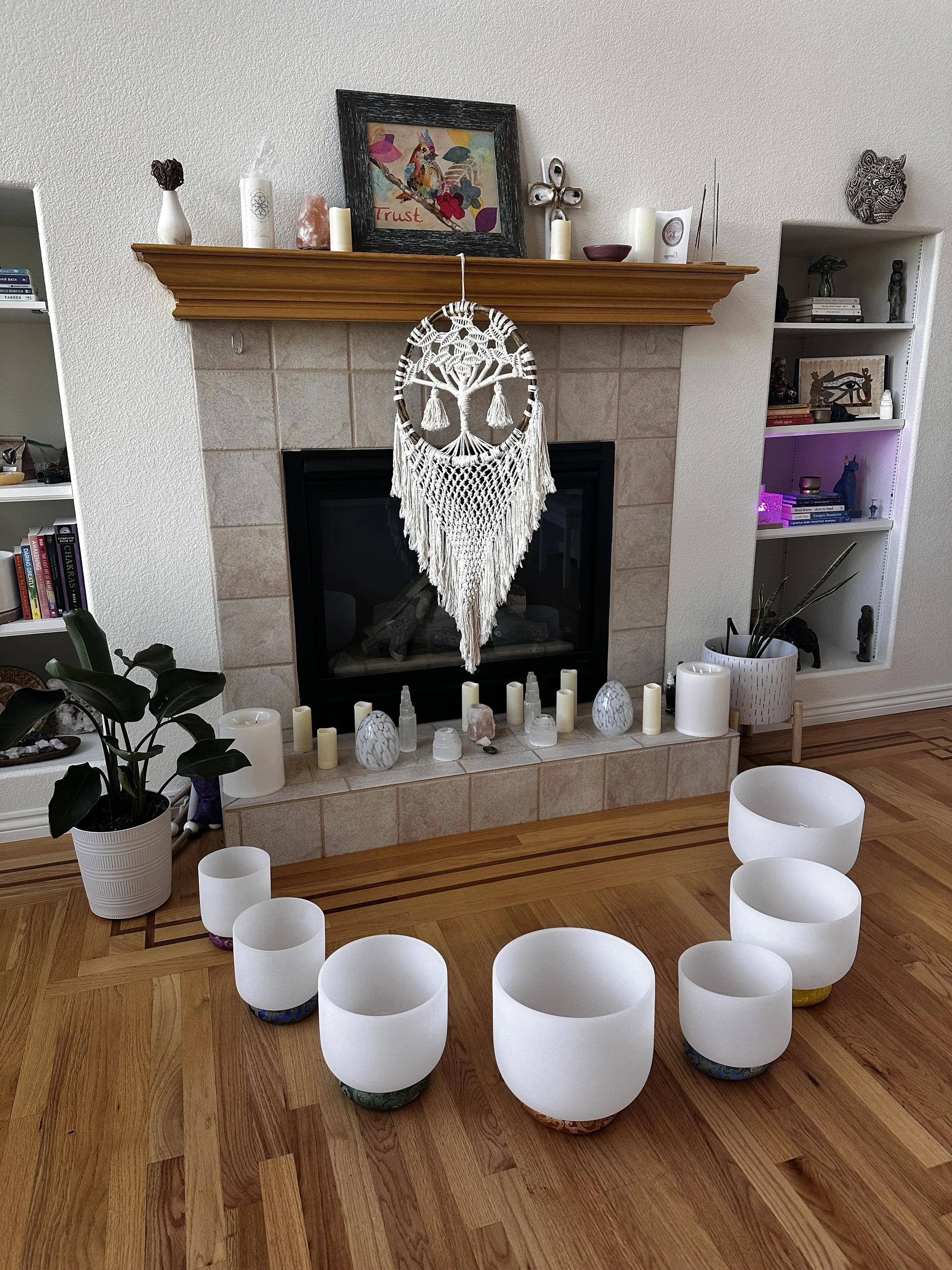 A living room fireplace decorated with candles, crystals, and a handmade macramé hanging in front of the fireplace. On the left, a potted plant, and on the right, a white bookshelf with books and decorative items. Multiple white cylindrical candles and bowls are placed on the floor in front of the fireplace. The room has hardwood flooring.