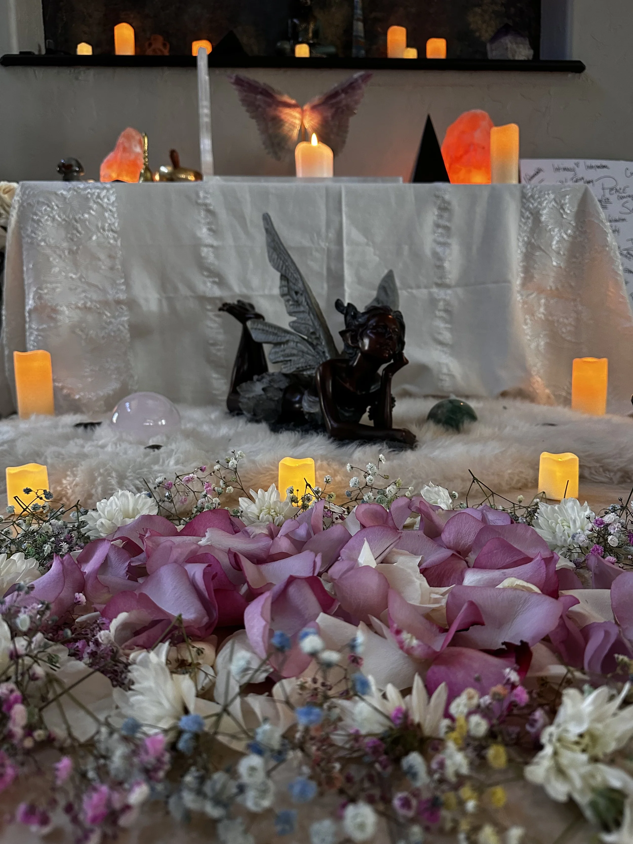 An altar adorned with pink and white flower petals, small flowers, and candles, with a central sculpture of a fairy with butterfly wings, candles, salt lamps, and a butterfly on the back wall.