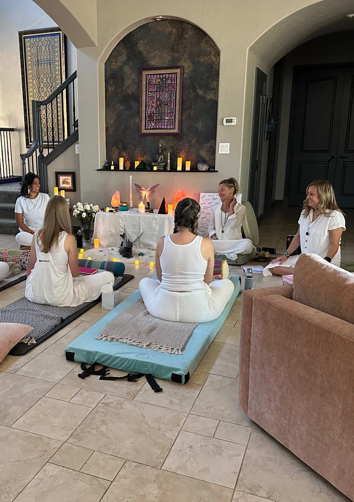 A group of women practicing yoga or meditation in a living room, seated on mats surrounded by candles and decorative items, with a shrine or altar in the background.
