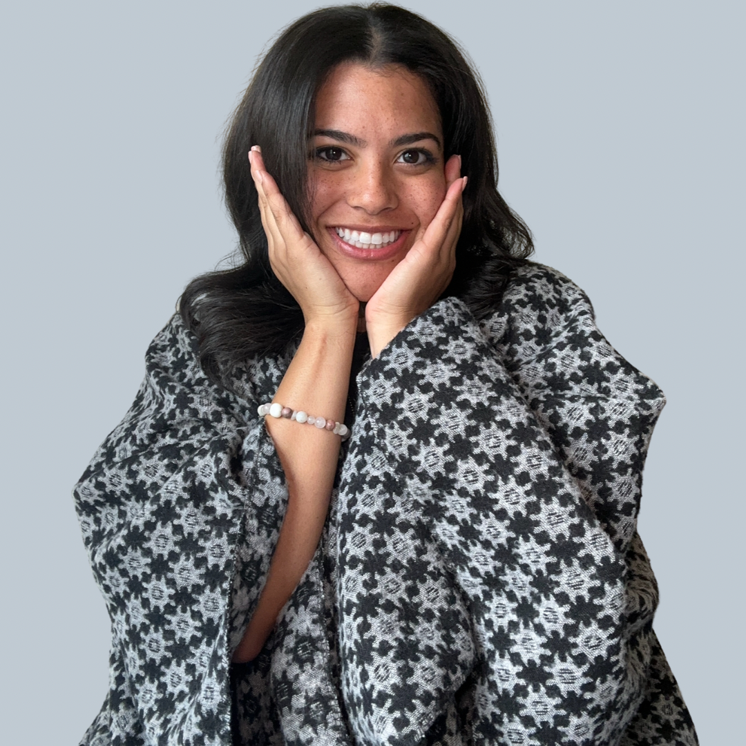 A woman with dark hair smiling, wearing a patterned black and gray top, holding her face with her hands against a plain light gray background.