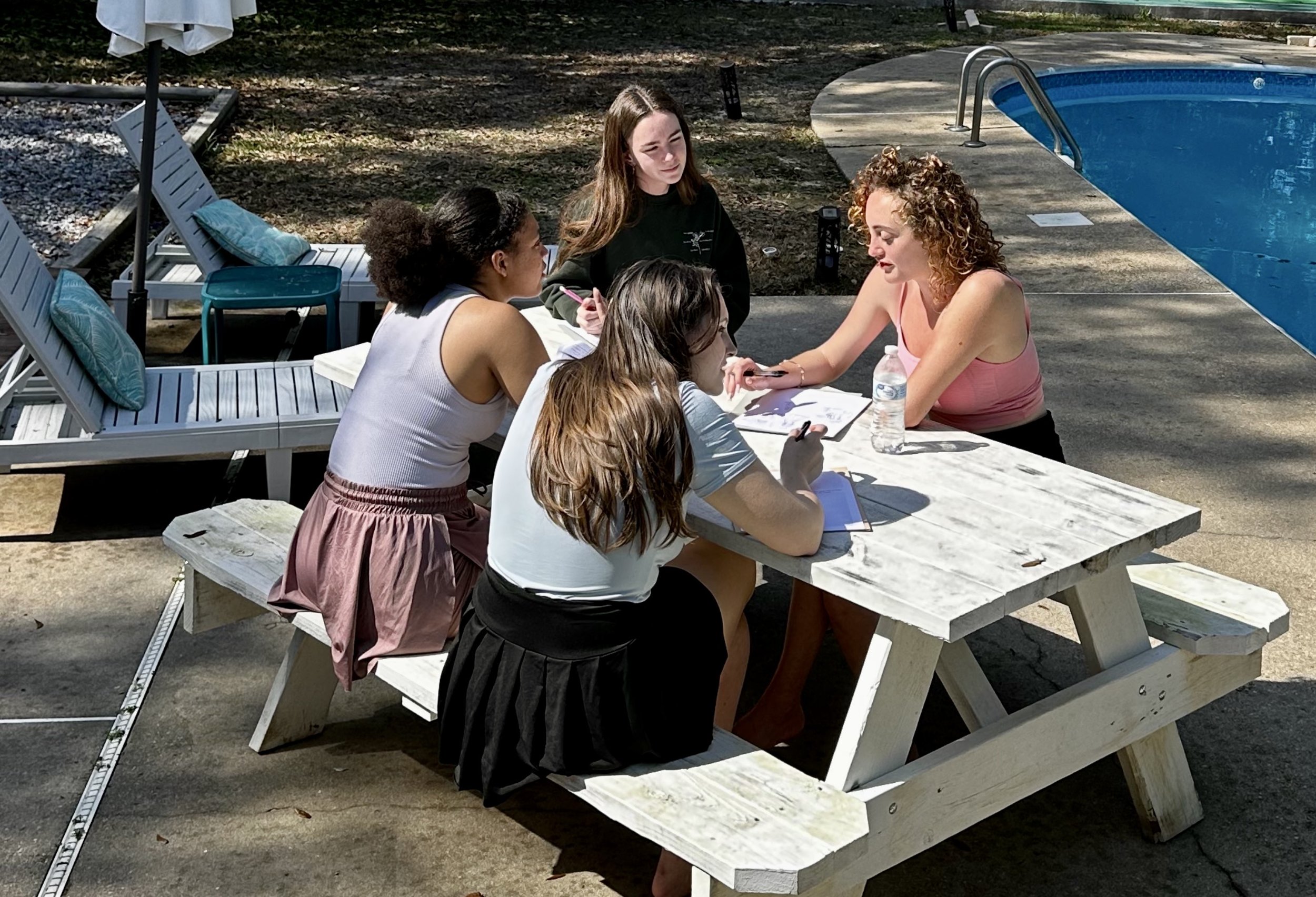 A group of five women sitting at a white wooden picnic table outdoors near a swimming pool, engaging in conversation or a meeting, with sunlight and shadows from nearby trees.