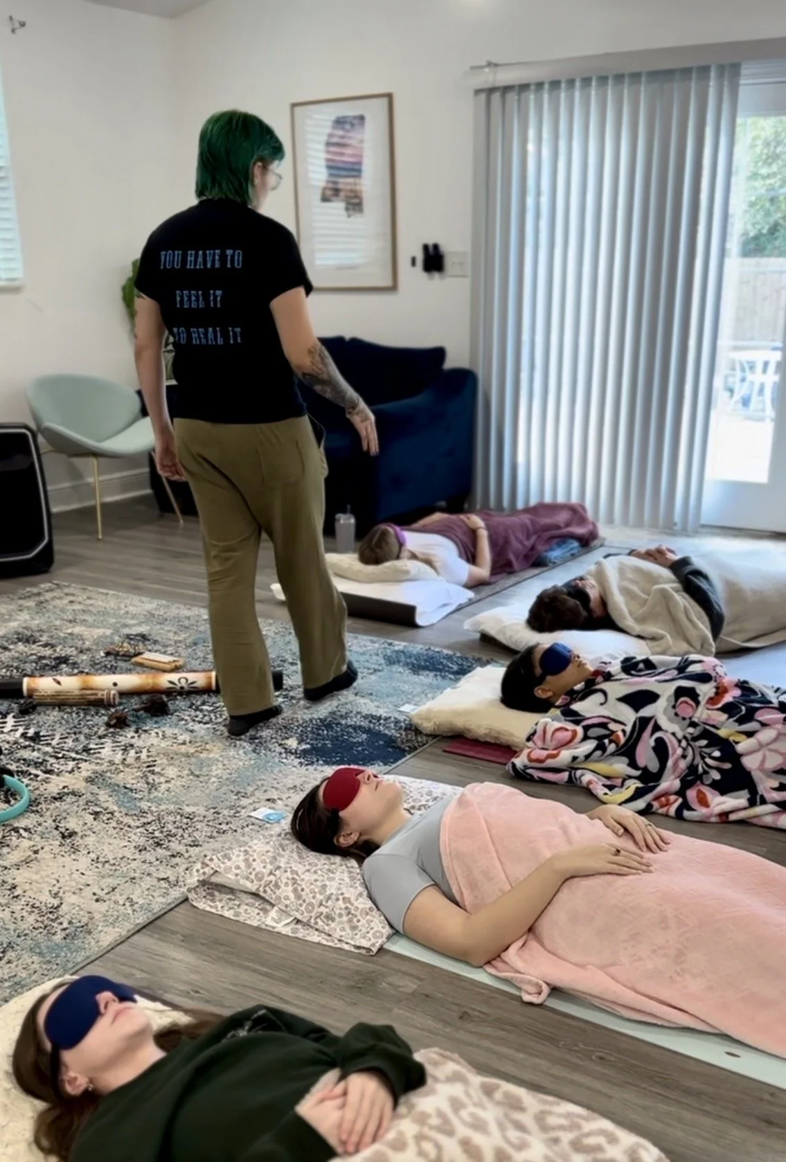 Multiple people are lying on the floor with eye masks, sleeping or resting. An instructor stands nearby in a living room setting with a sofa, artwork, and a sliding glass door.