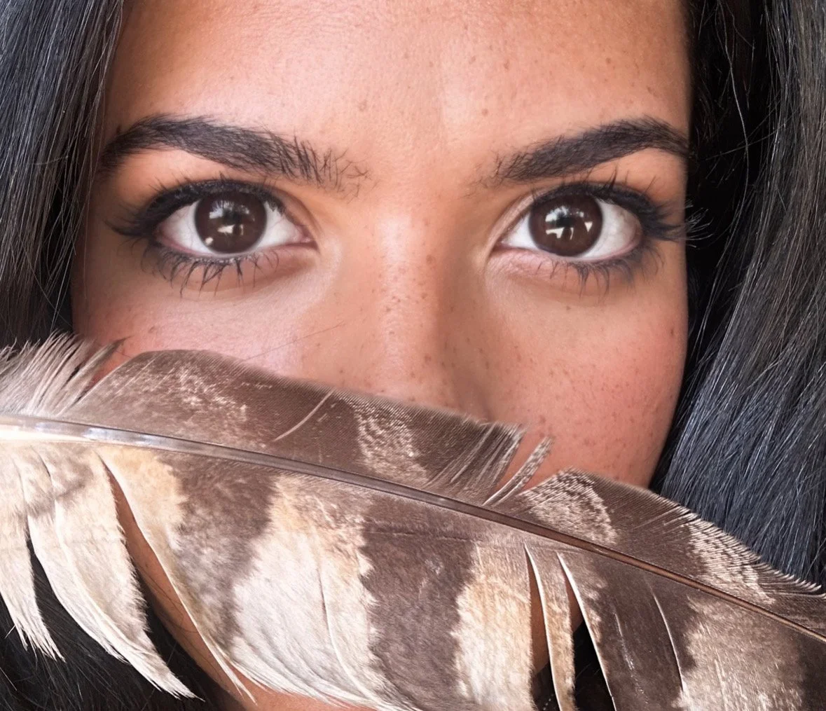 A close-up of a person's face, showing brown eyes, dark eyebrows, and dark hair, partially covered by a large feather.