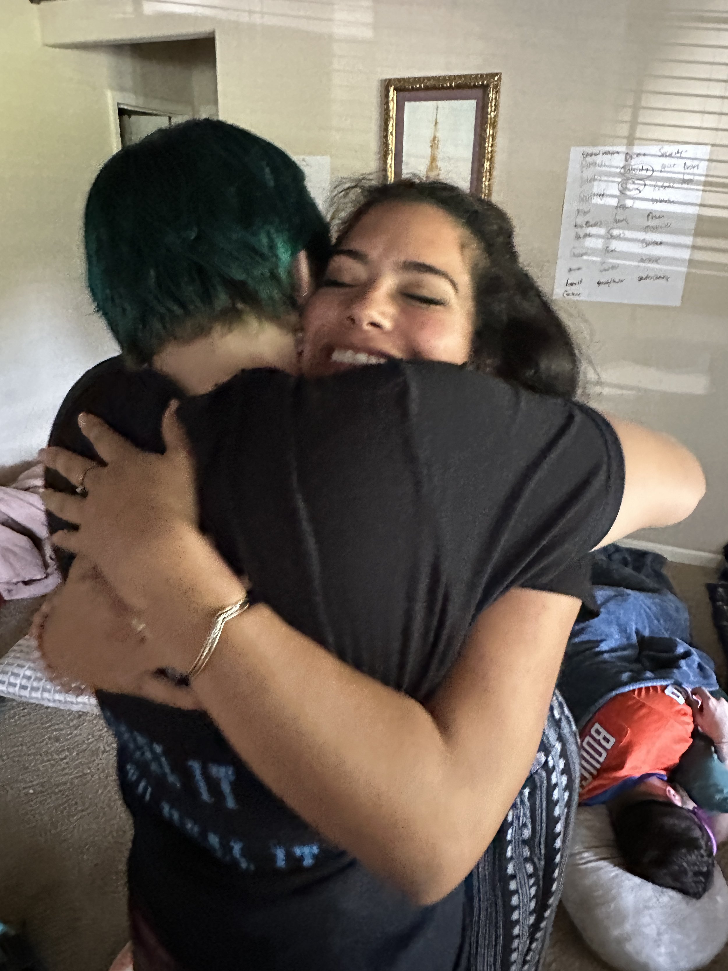 Two people embracing in a hug, smiling with eyes closed, in a room with a mirror and handwritten notes on the wall.