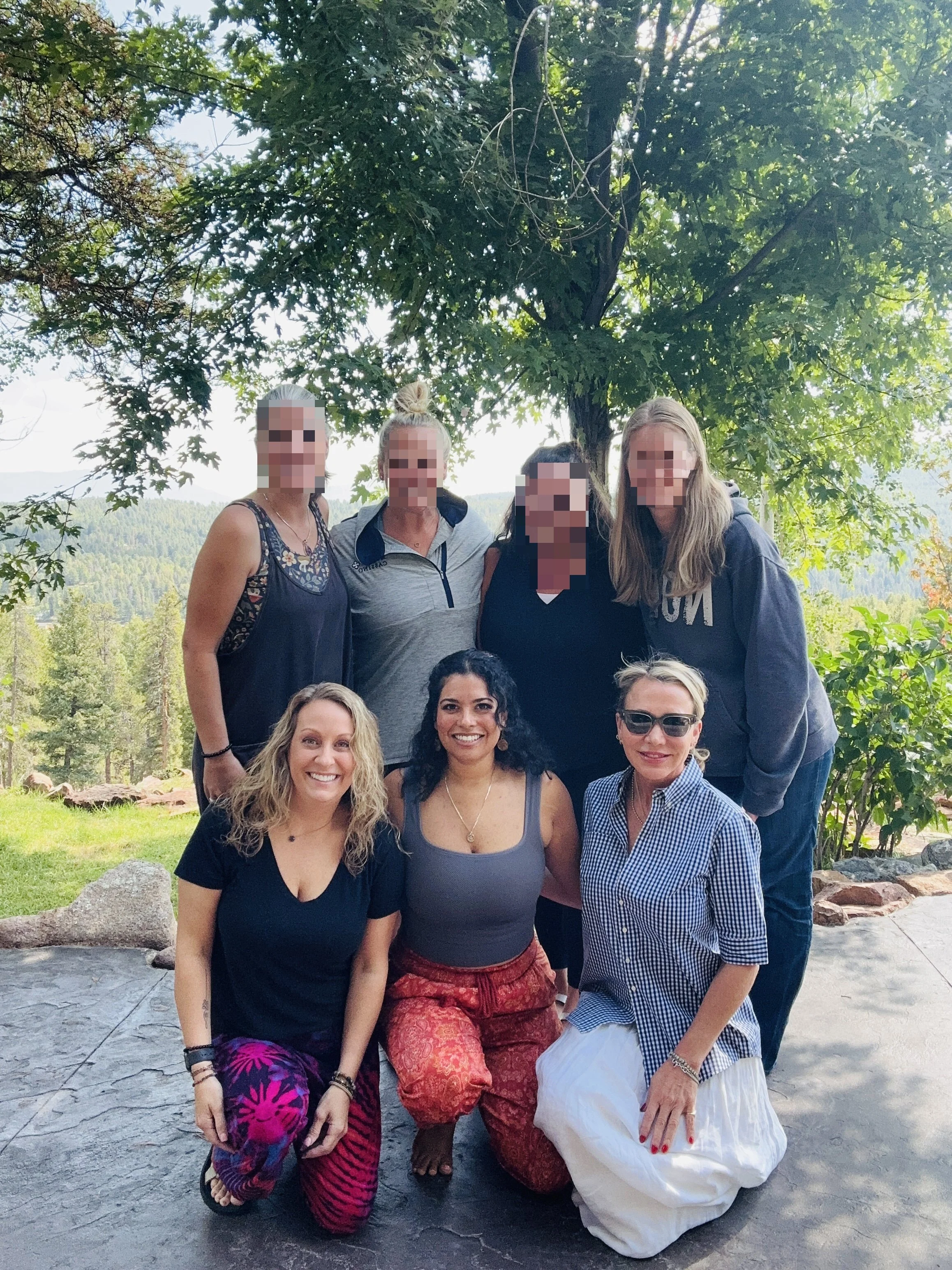 Group of nine women posing outdoors under a large tree with a scenic landscape in the background, some standing, some kneeling, dressed in casual summer clothing.