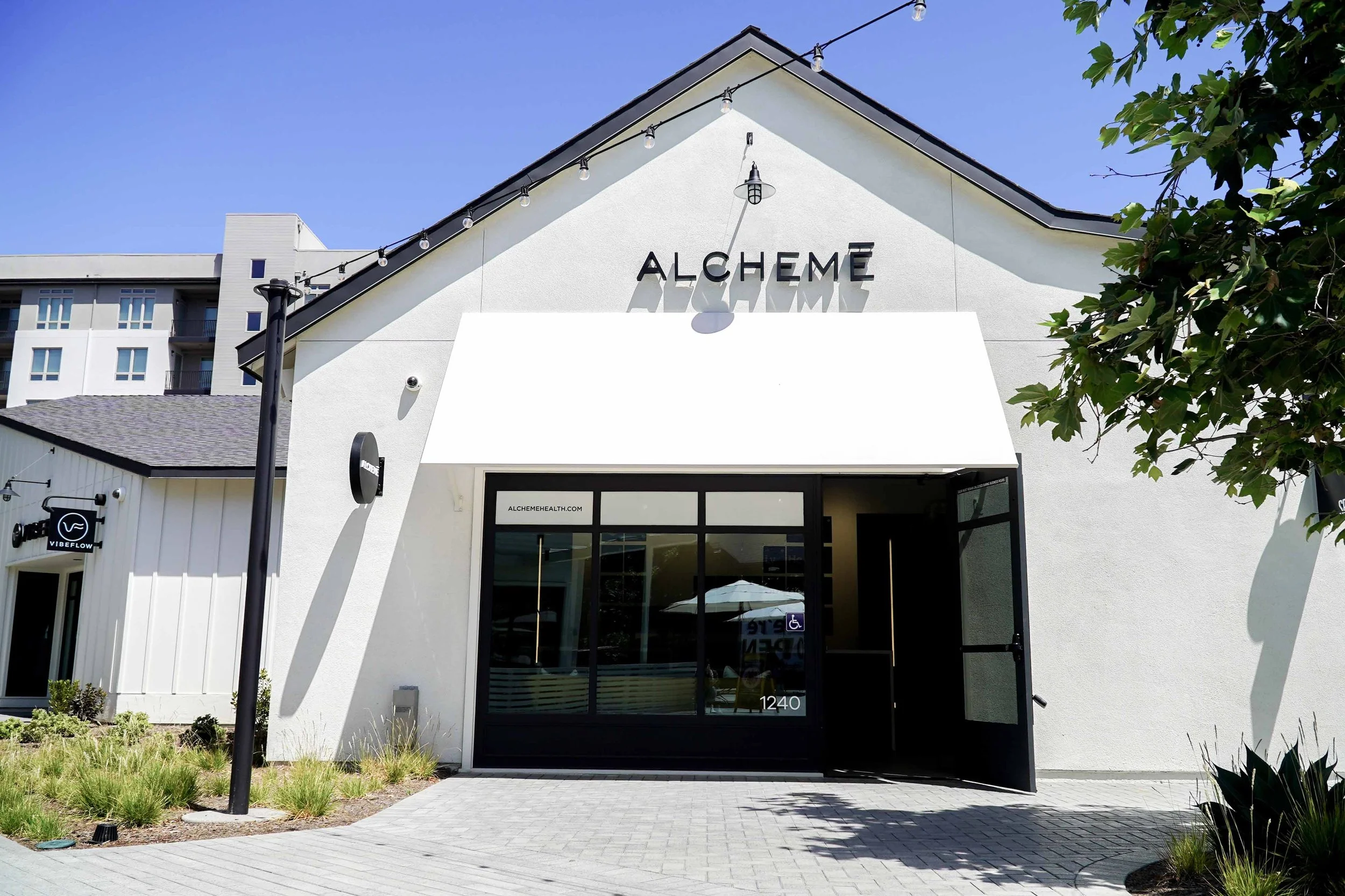 Exterior of a modern building with "ALCHEME" signage, featuring a white façade, black trim, and a large awning. A sign with the website "alchemehealth.com" is on the glass door. Nearby, another building's sign reads "VIBE FLOW."