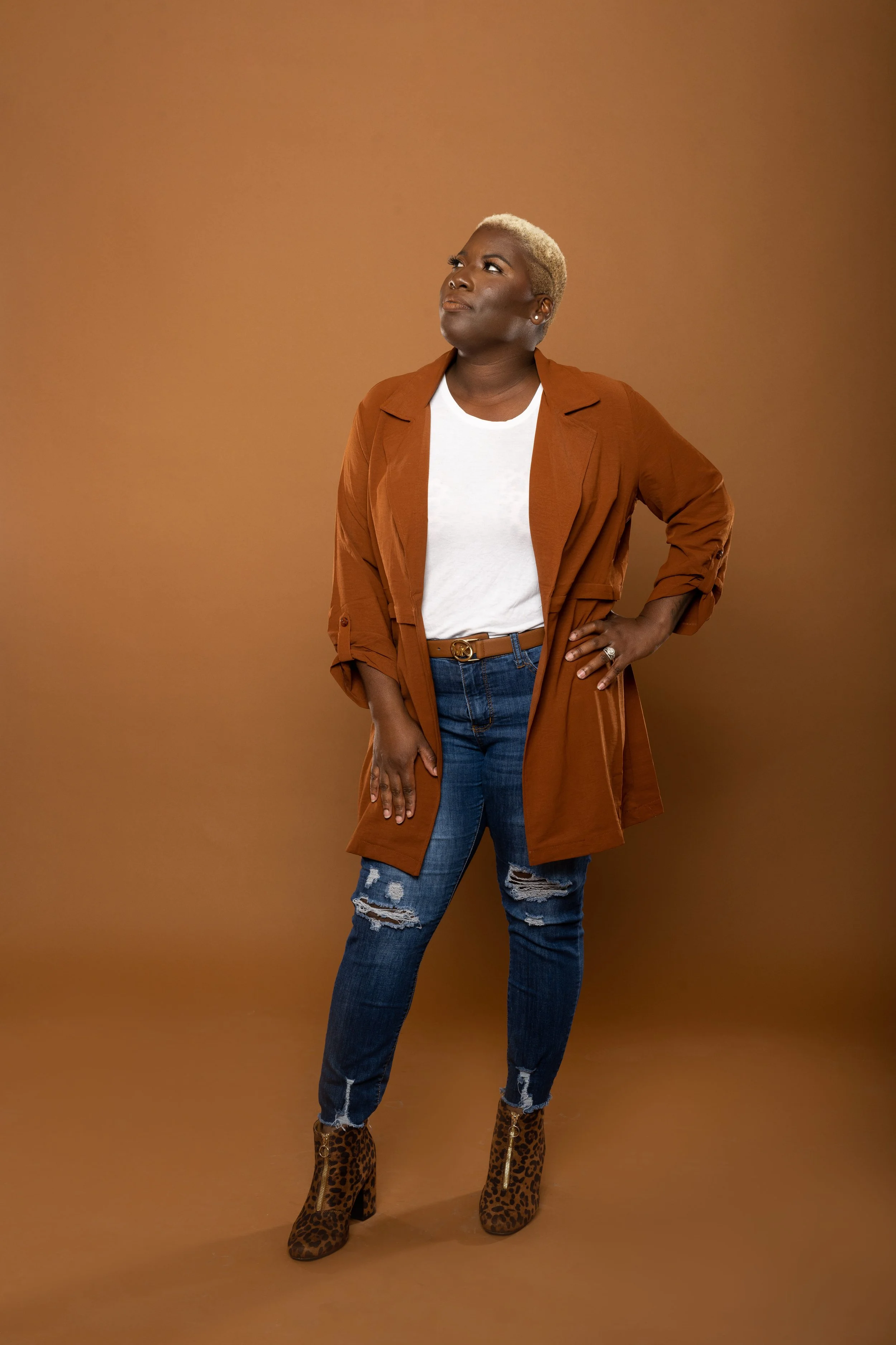 A woman with short blonde hair wearing a white t-shirt, brown trench coat, ripped blue jeans, and leopard print high-heeled boots stands against a tan background.