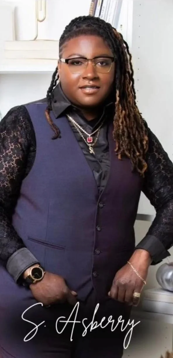 A woman with glasses, wearing a dark purple vest over a black lace long-sleeve top, accessorized with jewelry, standing indoors with a white shelf and books behind her, and the text 'S. Asbury' at the bottom.