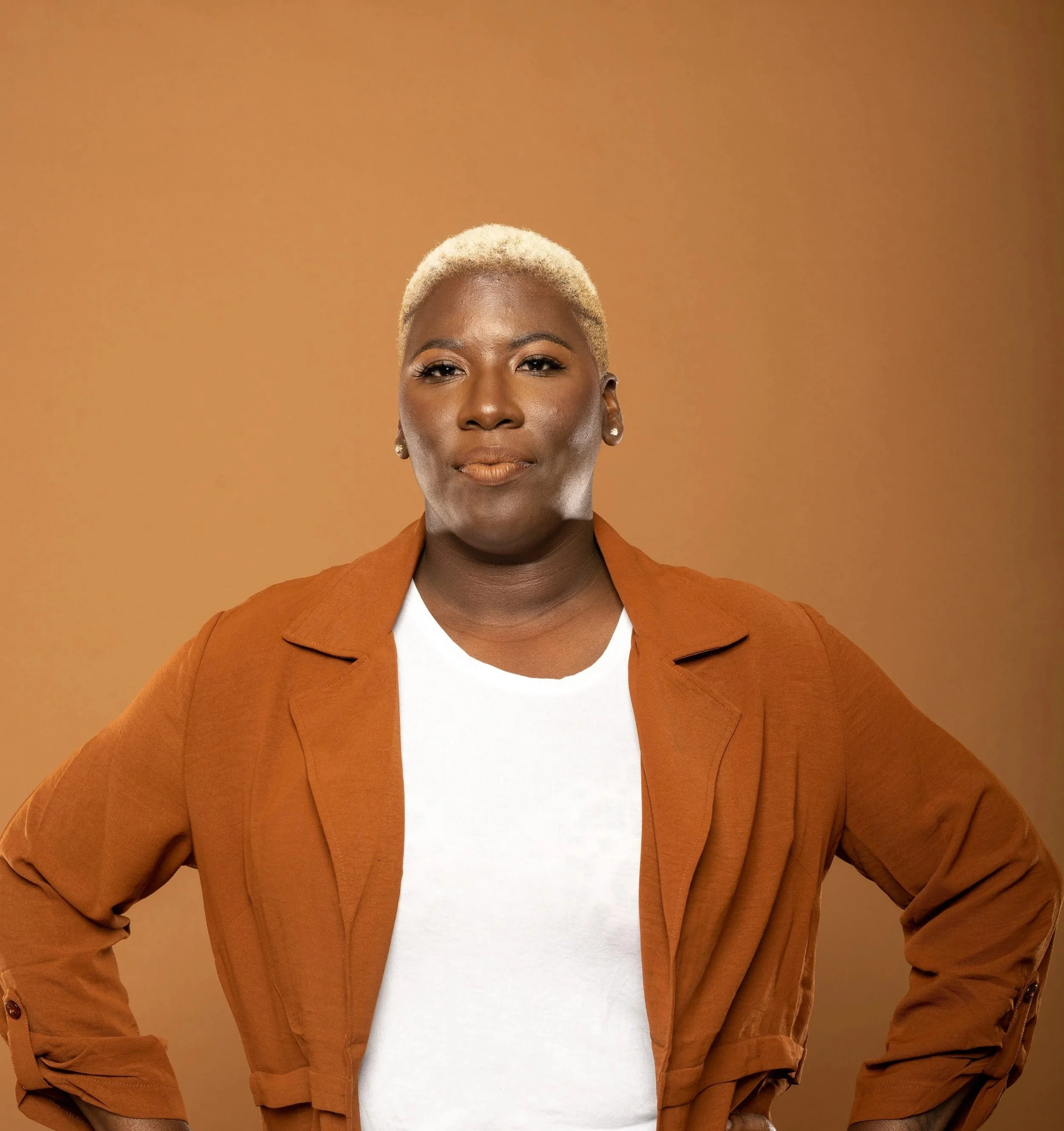 A confident woman with short blonde hair, wearing a white shirt, a brown belt, blue jeans, and a brown coat, standing with hands on hips against a brown background.