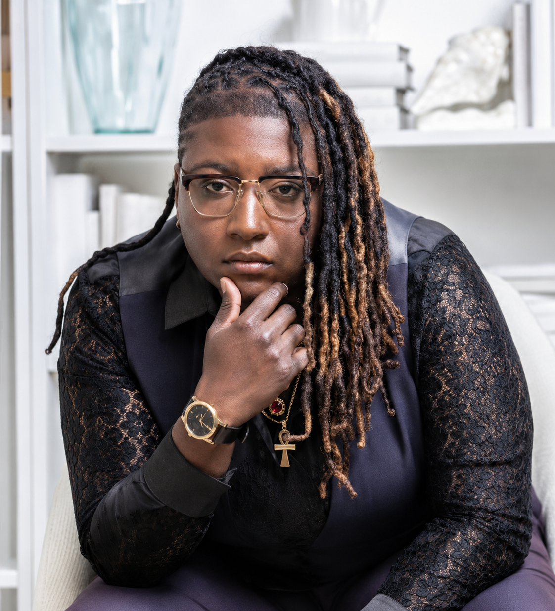 A woman with glasses and dreadlocks resting her chin on her hand, wearing a black lace top and a watch, sitting in a room with white bookshelves in the background.