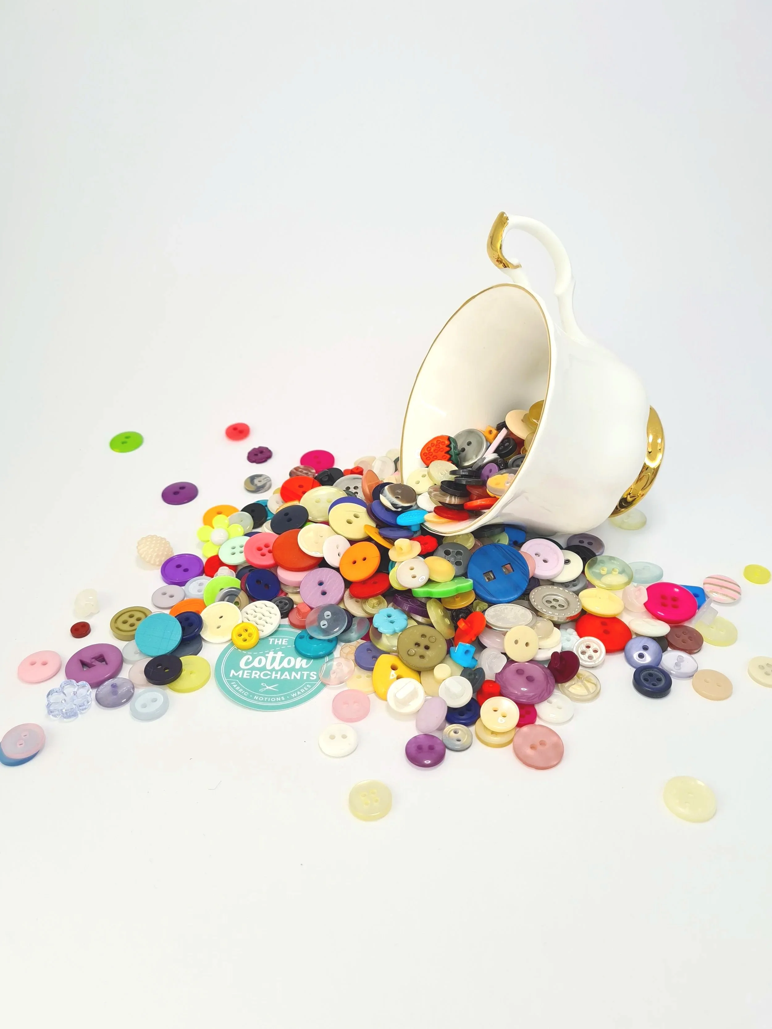 A white teacup tipped over on a white surface, spilling a variety of colorful buttons of different sizes and shapes, with some scattered around the cup.