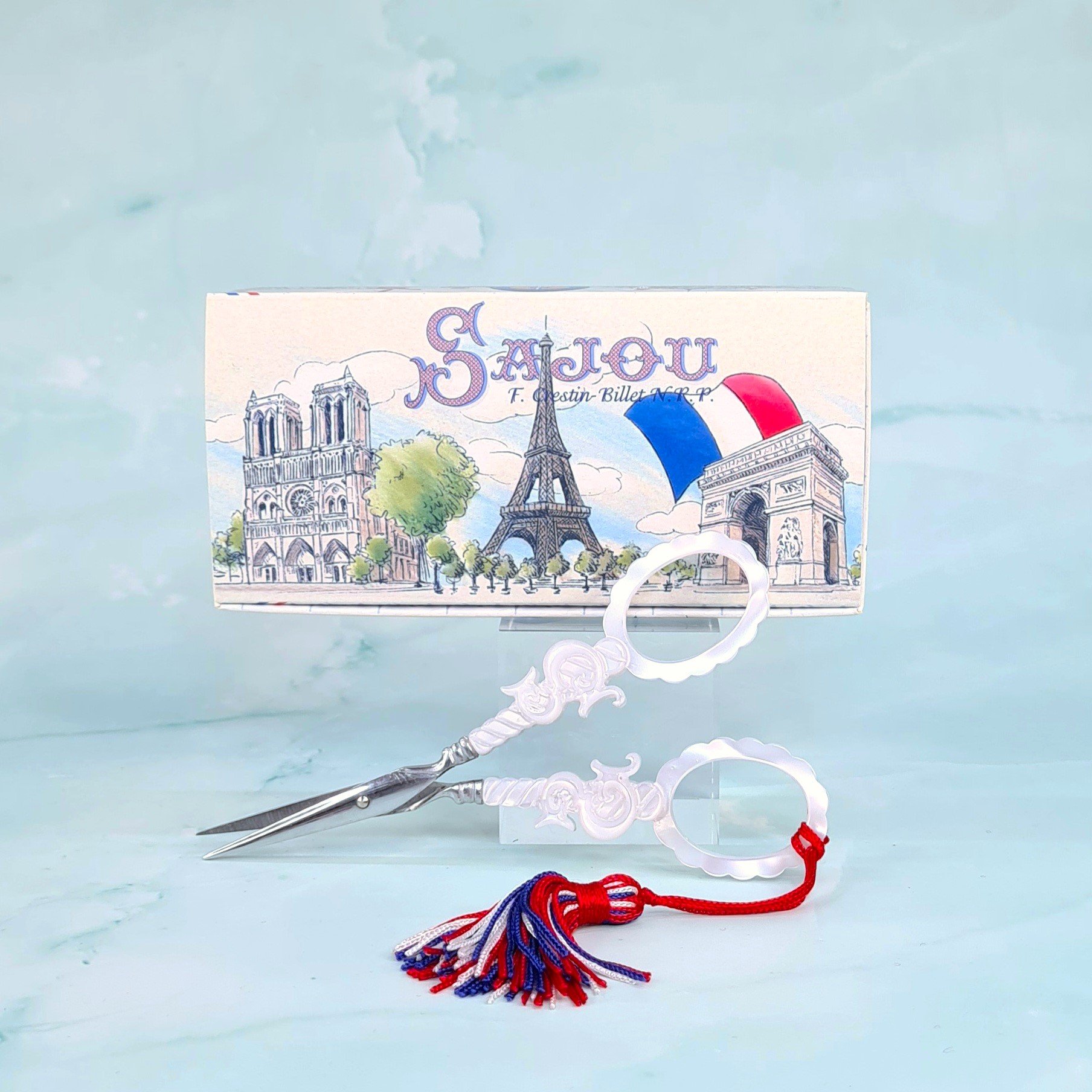 A Paris-themed souvenir box with a pair of pearl-handled scissors, featuring illustrations of Notre-Dame Cathedral, the Eiffel Tower, and the Arc de Triomphe on the box, set against a light blue background.
