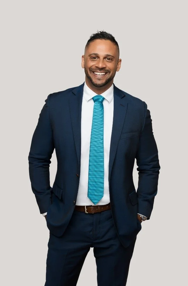 A smiling man in a navy blue suit, white shirt, and matching blue tie, standing against a plain light gray background.