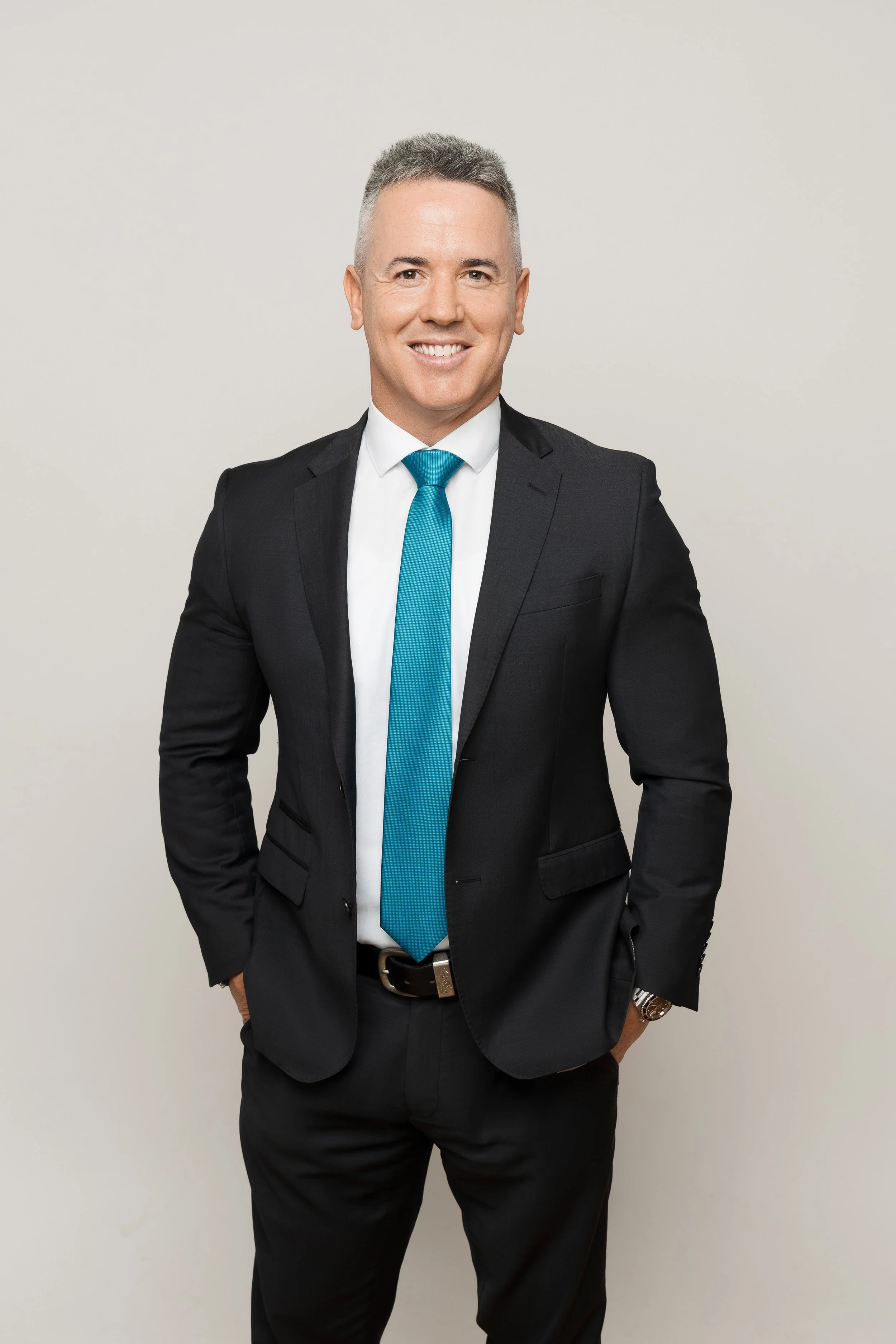 A man in a black suit, white shirt, and teal tie standing against a plain light-colored background.